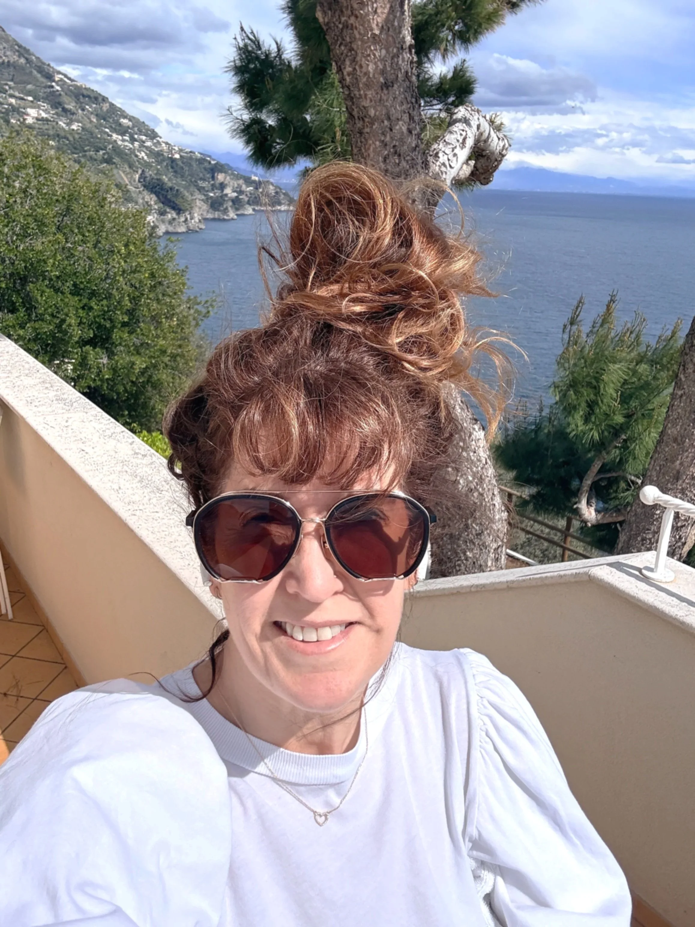 A woman with curly reddish-brown hair and large sunglasses taking a selfie outdoors on a balcony overlooking the ocean, with mountains and cloudy sky in the background.