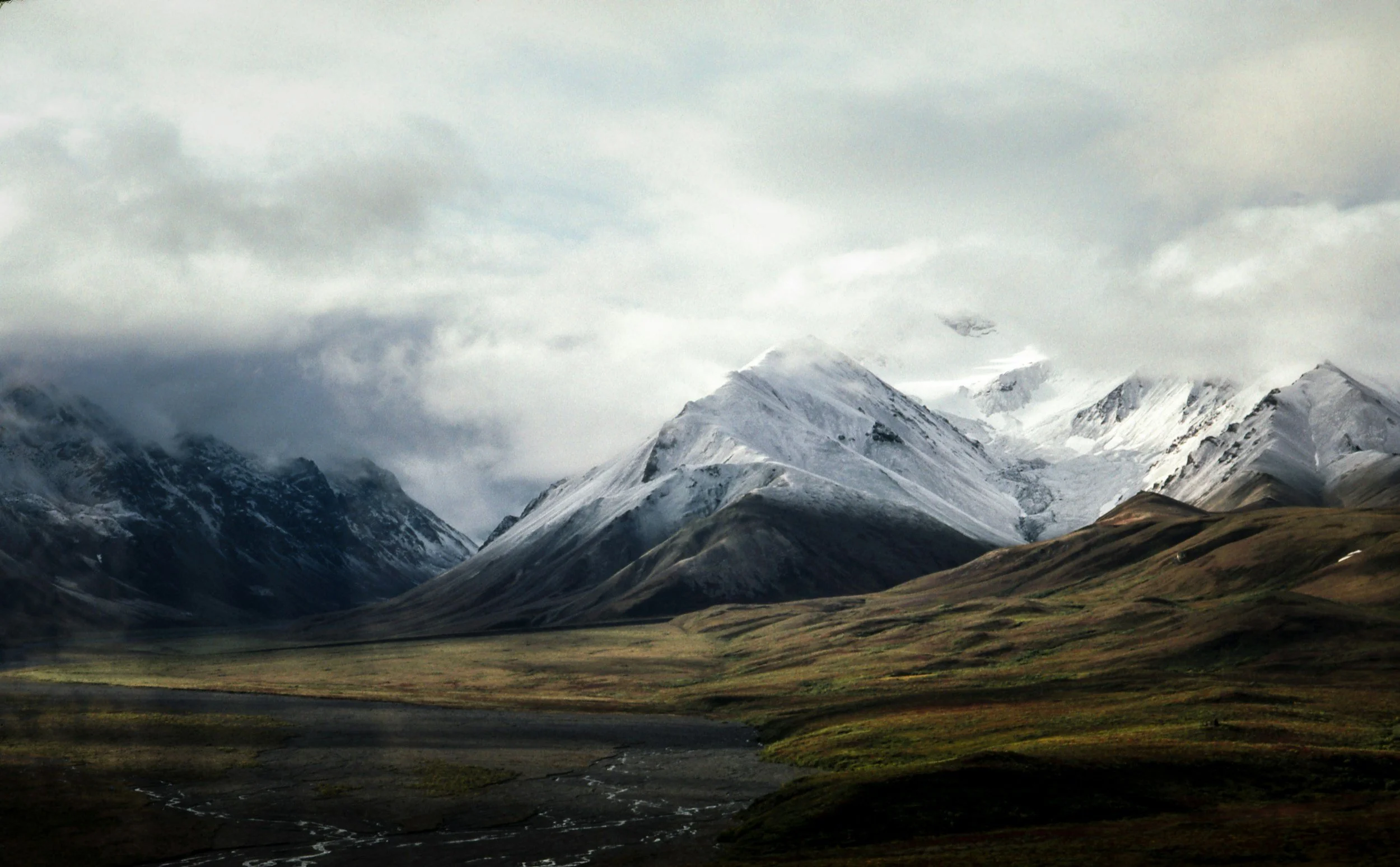 Snow-capped mountains under cloudy sky with green rolling hills at the base.