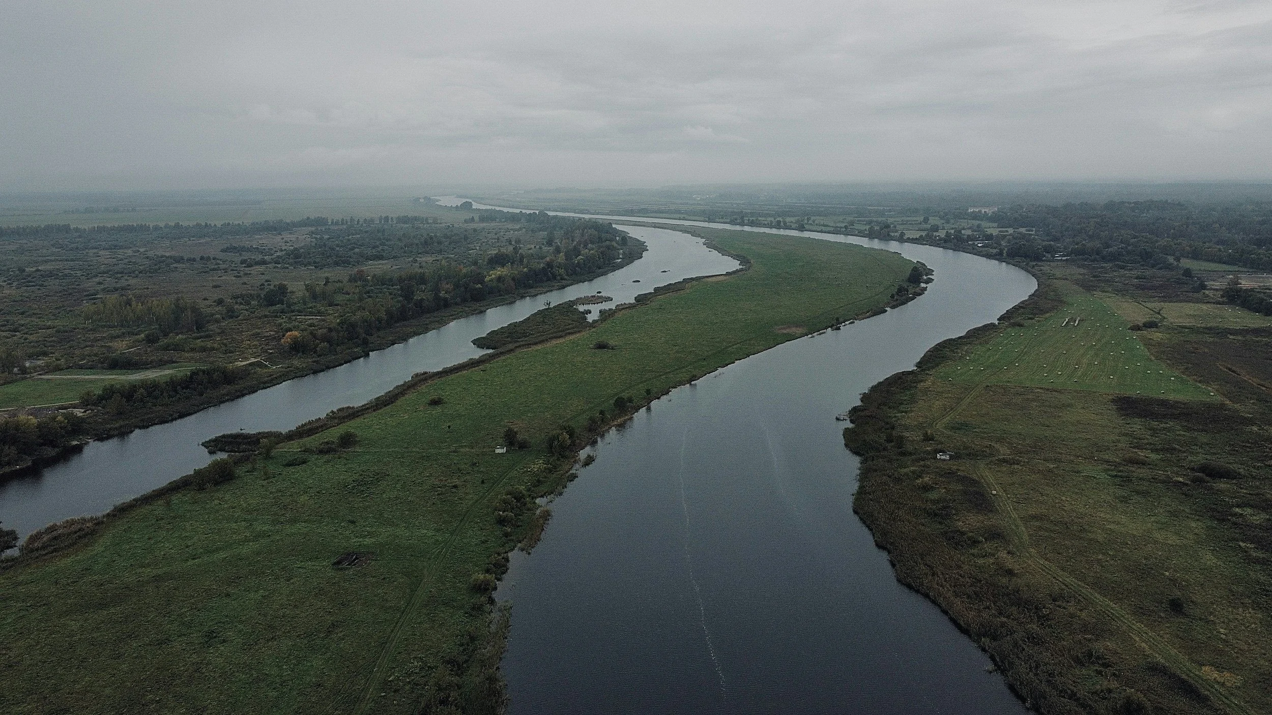 Aerial view of a winding river flowing through green fields and distant farmland under gray cloudy skies.