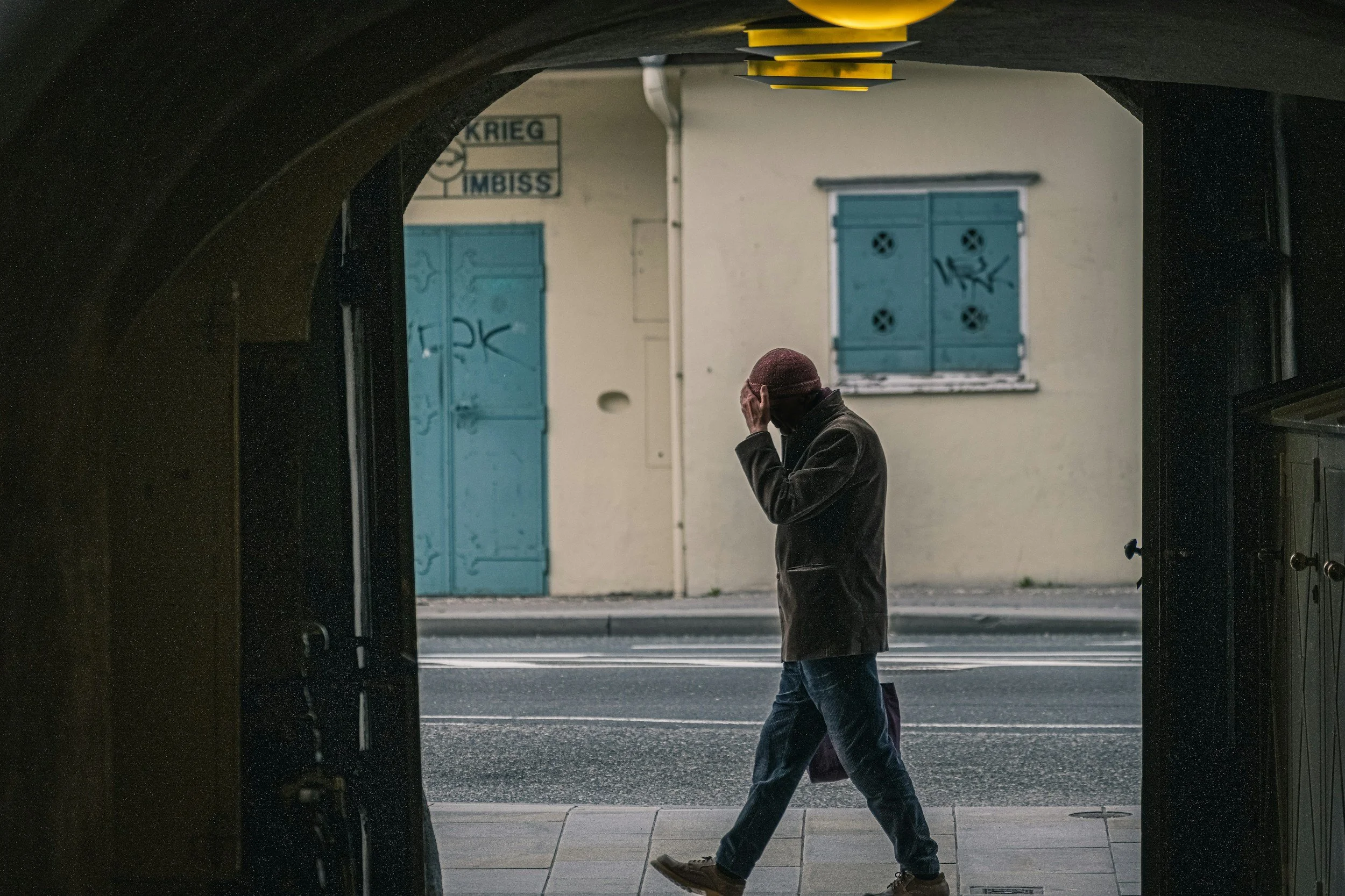 A person walking past a building with blue shutters, as seen through a dark archway. The person is wearing a red hat, a dark jacket, and jeans, and appears to be touching their head with one hand.
