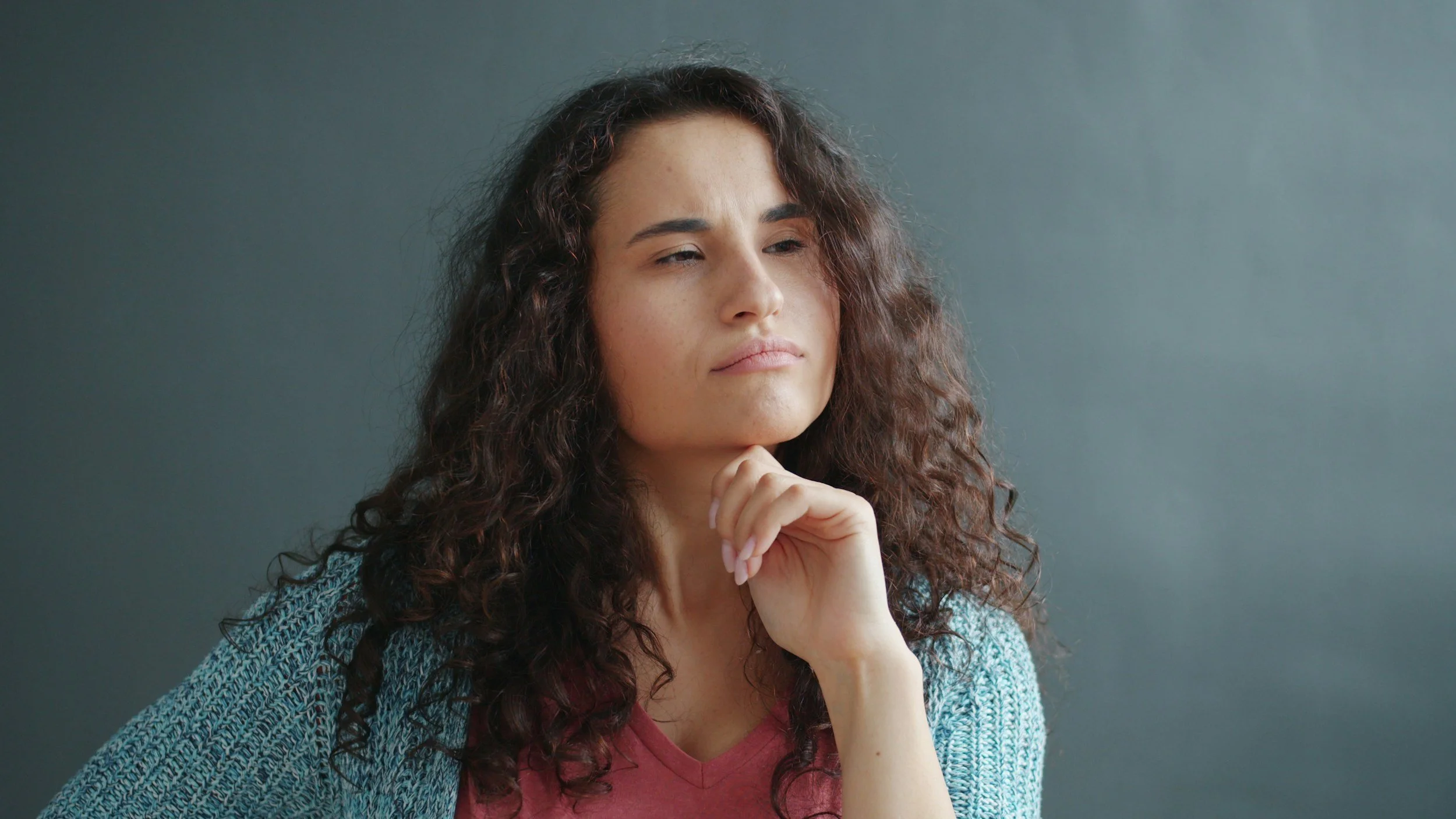 A woman with curly brown hair, wearing a blue sweater over a maroon top, is resting her chin on her hand and looking thoughtfully to the side.