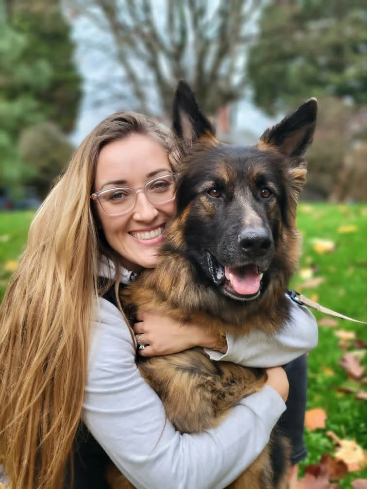A smiling woman with long blonde hair, glasses, and a white jacket is smiling and hugging a large German Shepherd dog outdoors in a park with green grass and trees.