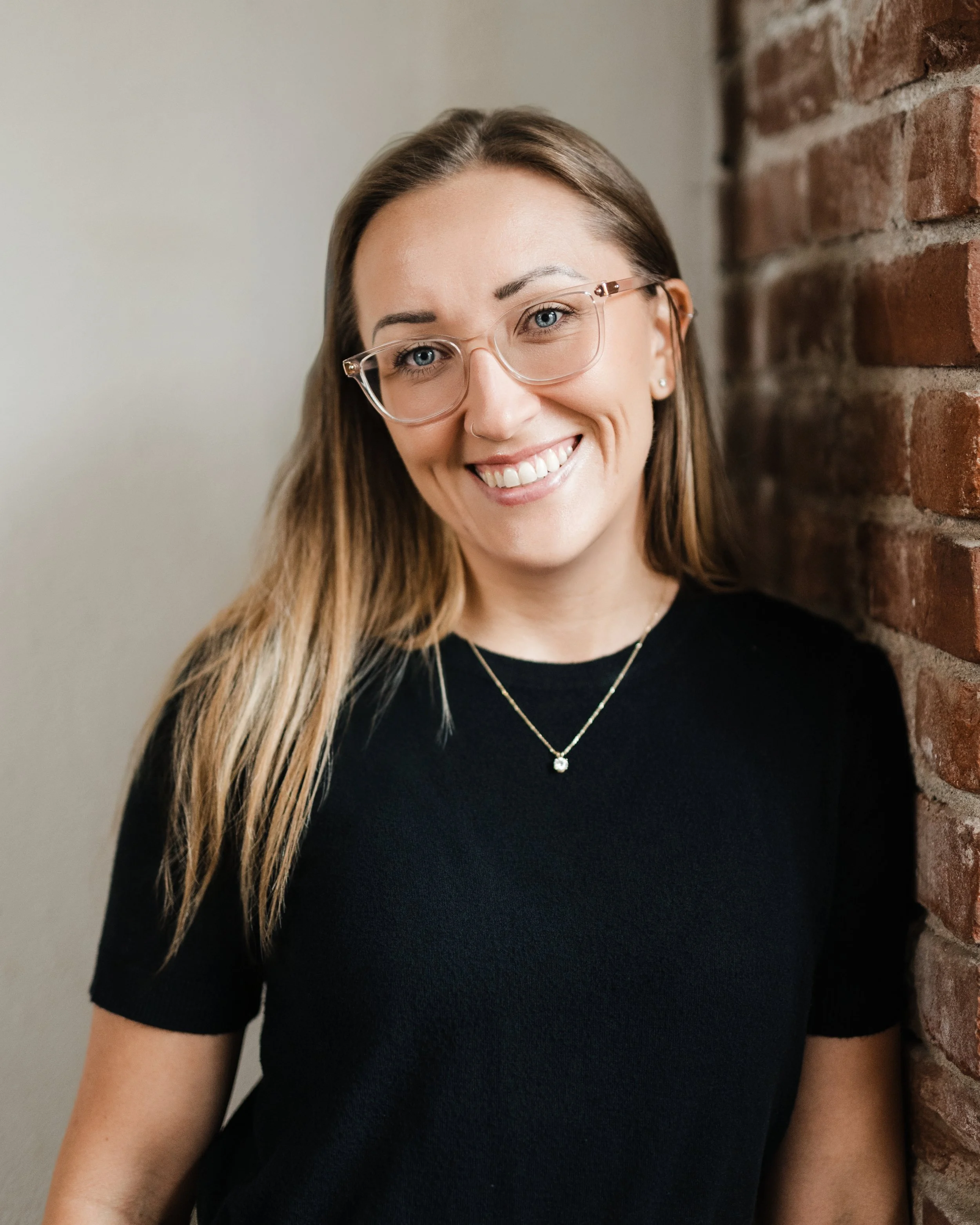 A woman with long brown hair, wearing glasses, a black shirt, and jewelry, smiling while leaning against a brick wall.