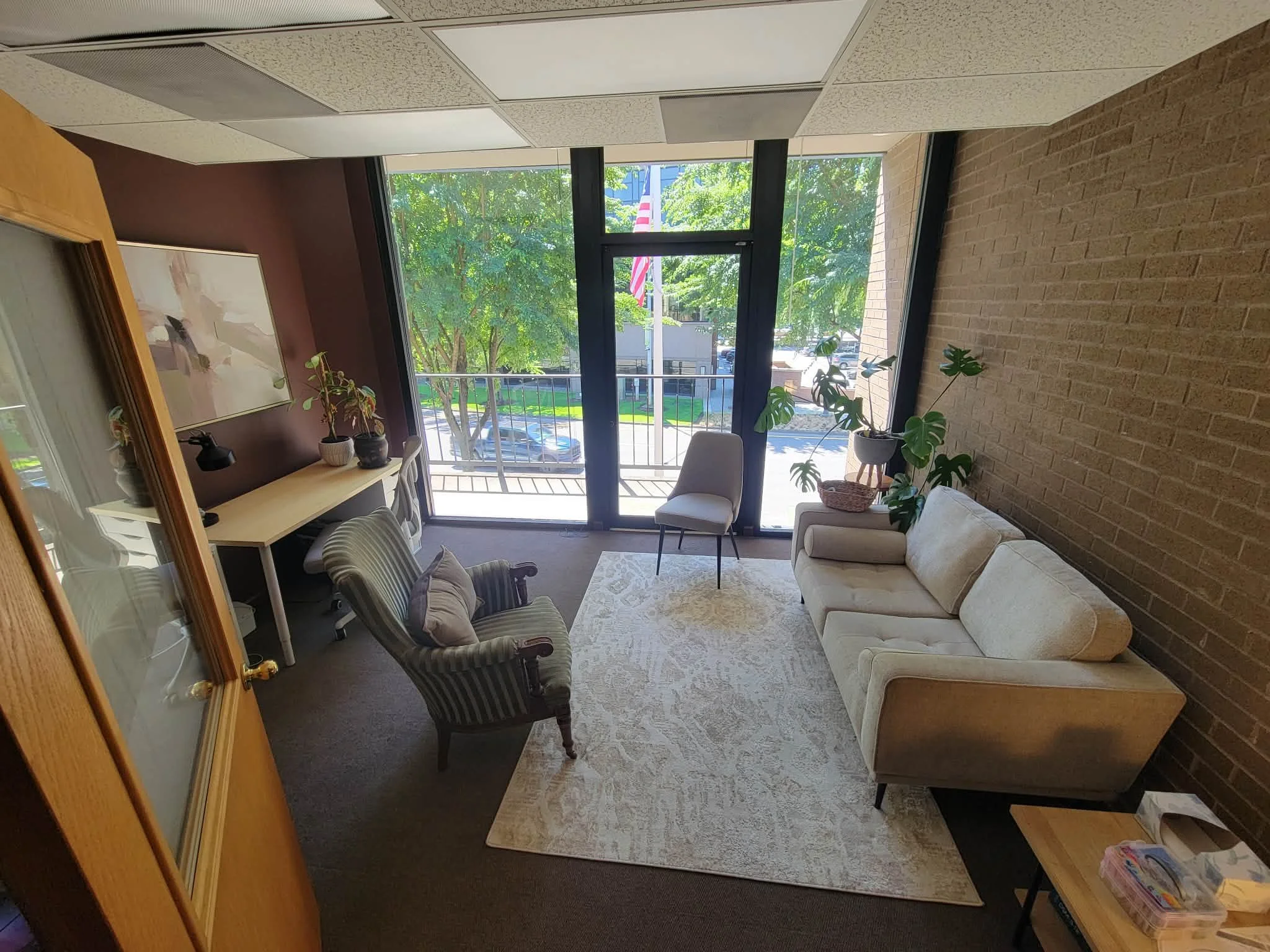A cozy living room with a large glass door leading to a balcony with trees outside. The room has a beige sofa, a striped armchair, and several potted plants. There is a bookshelf with decorative items and a painting on a dark accent wall.