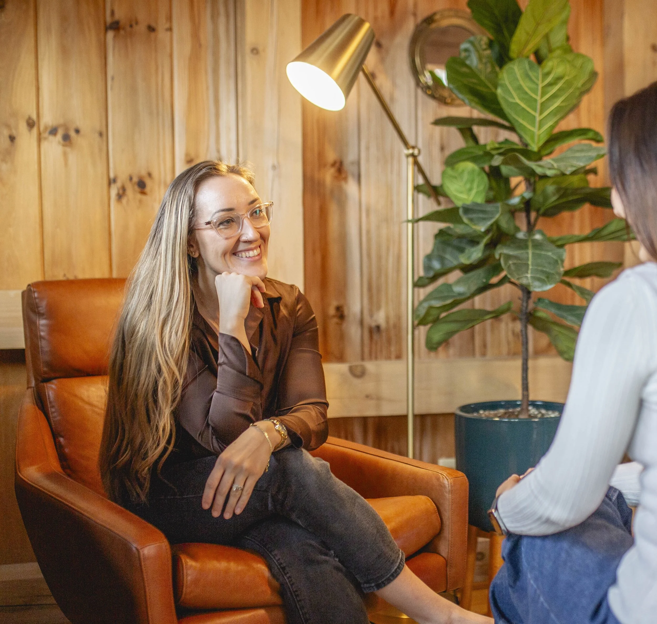 A woman with long blonde hair, glasses, and a brown shirt sitting on an orange leather armchair, smiling and having a conversation with another person in a cozy wooden room with a large potted plant and a floor lamp.