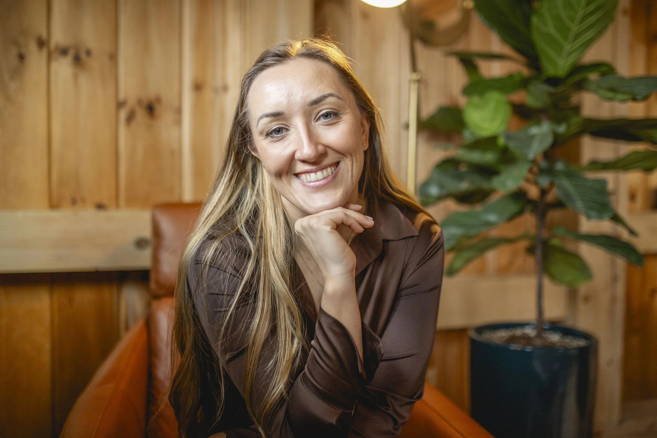 A woman with long blonde hair smiling, sitting on a wooden bench with a wooden wall and a large green leafy plant in the background.