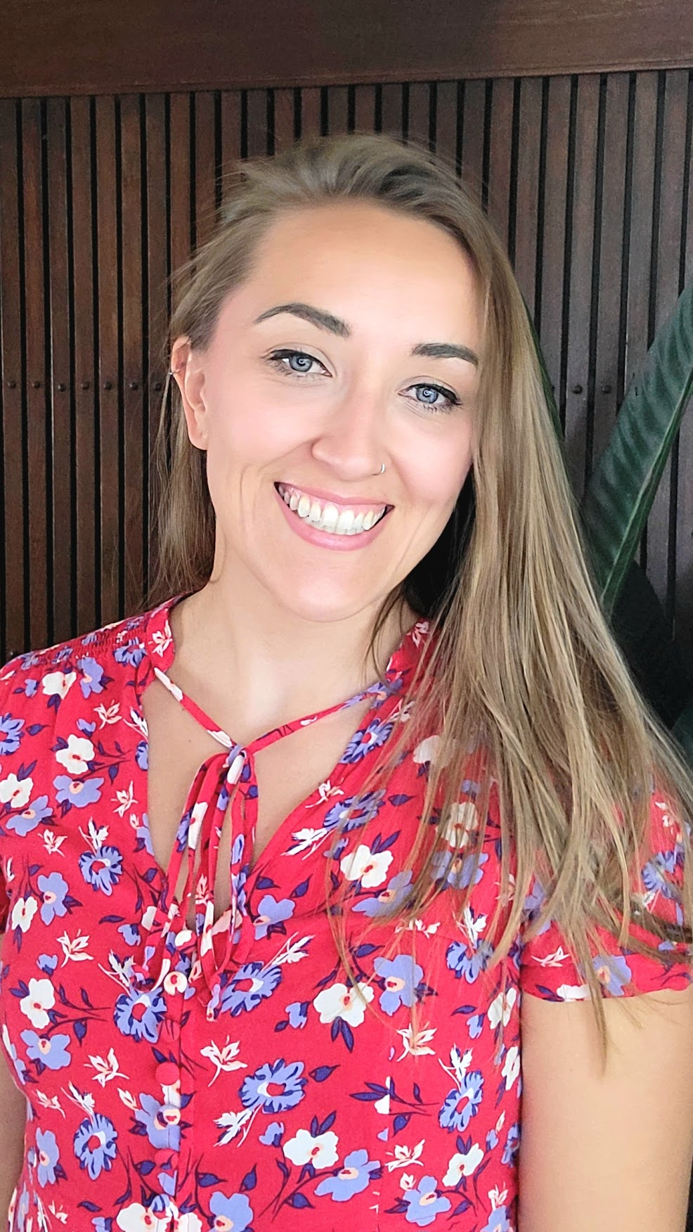 A smiling woman with long, light brown hair wearing a red floral top with a bow, standing against a wooden wall with vertical slats and a green plant.