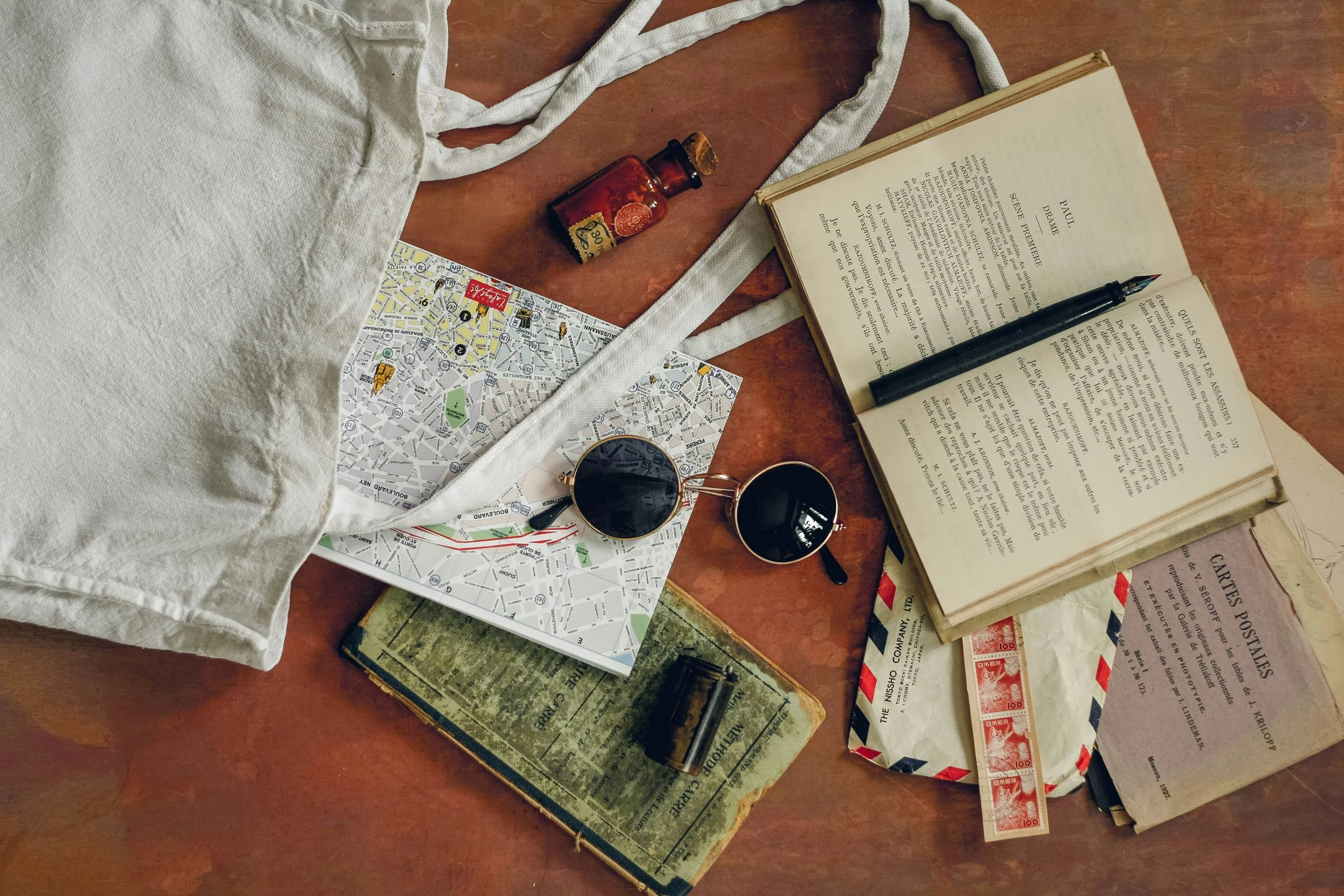 a map, pair of sunglasses, open book, amber colored glass vial with cork, small canister, and canvas bag lay on a wooden table.