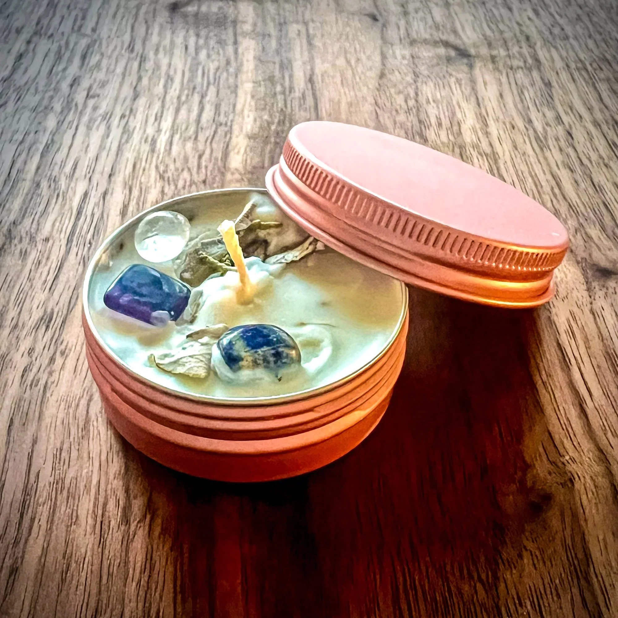 A mini self-love ritual candle in pink tin container with lid rests on a wooden surface. These hand poured, small batch ritual candles are infused with blessed intentions, topped with sacred herbs and crystals.