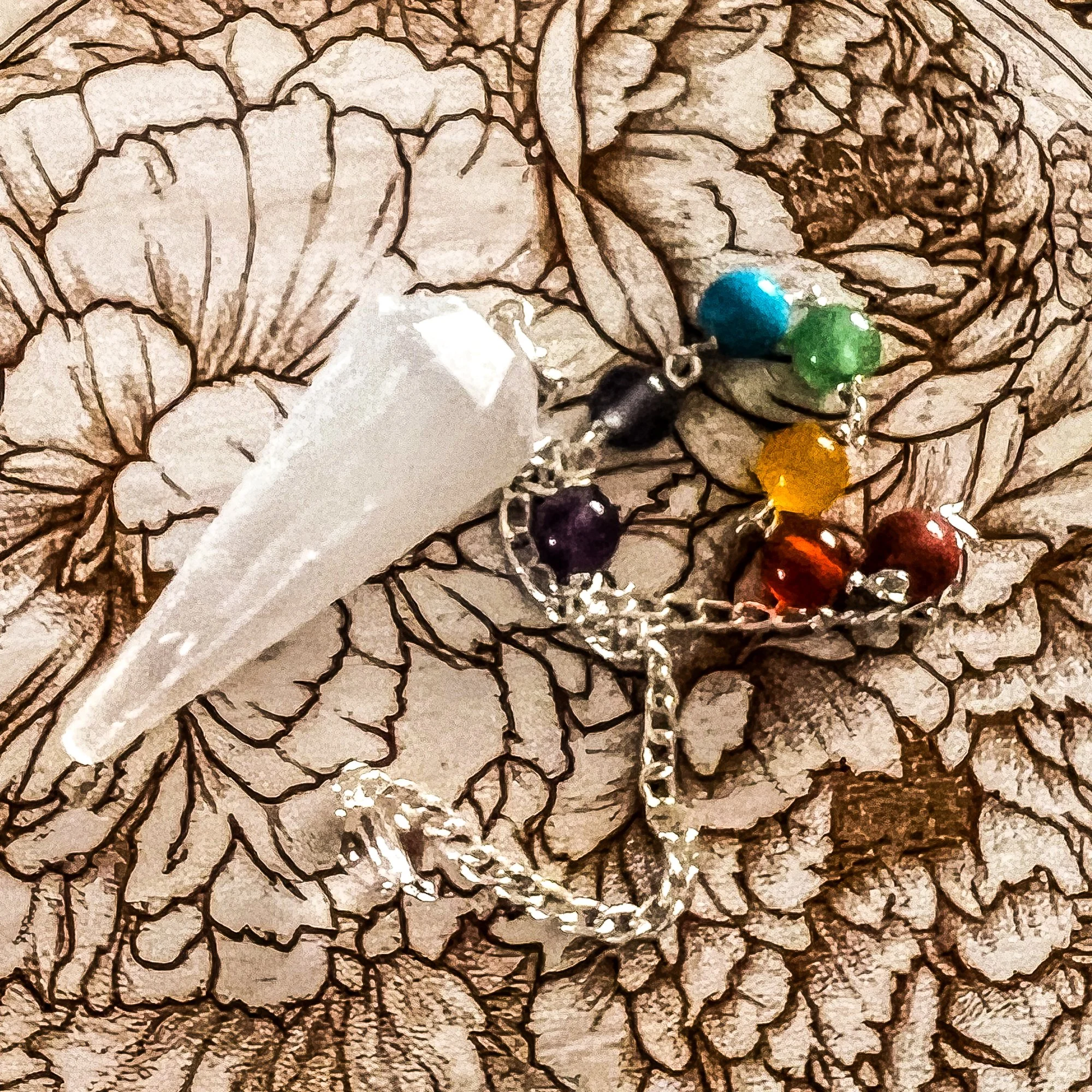 selenite scrying pendulum with semi-precious stones in chakra-aligned colors on chain, resting on hand-painted engraved chrysanthemum pendulum board