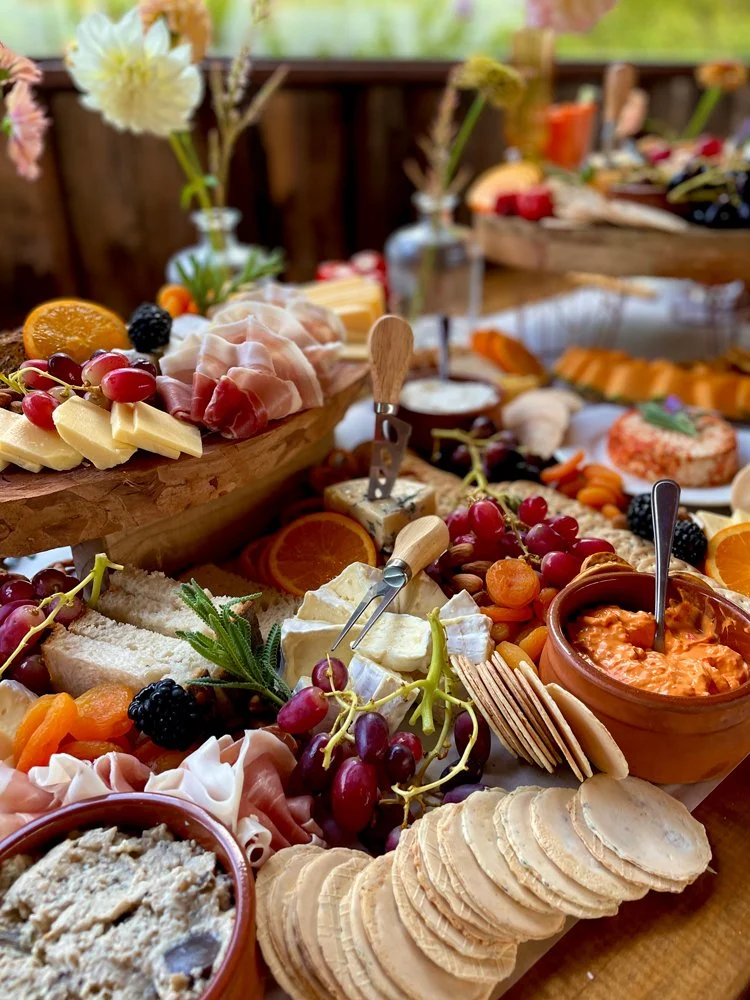 Assorted cheeses, crackers, grapes, cherry tomatoes, sliced meats, and dips arranged on a wooden table for a charcuterie board, with flowers in vases in the background.