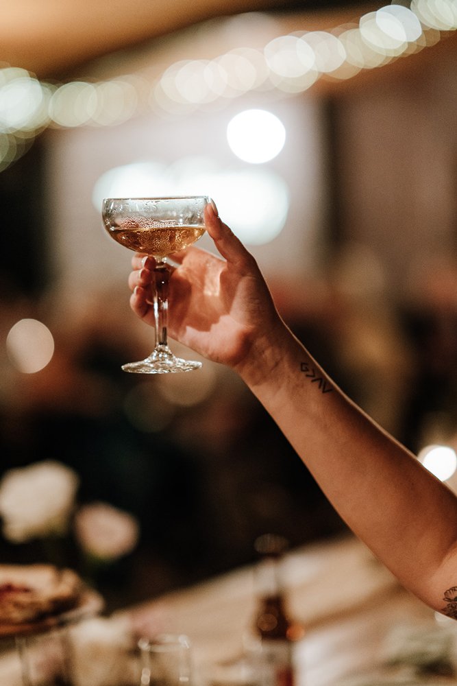 A hand holding a champagne coupe filled with sparkling wine or champagne, with blurred party lights in the background.