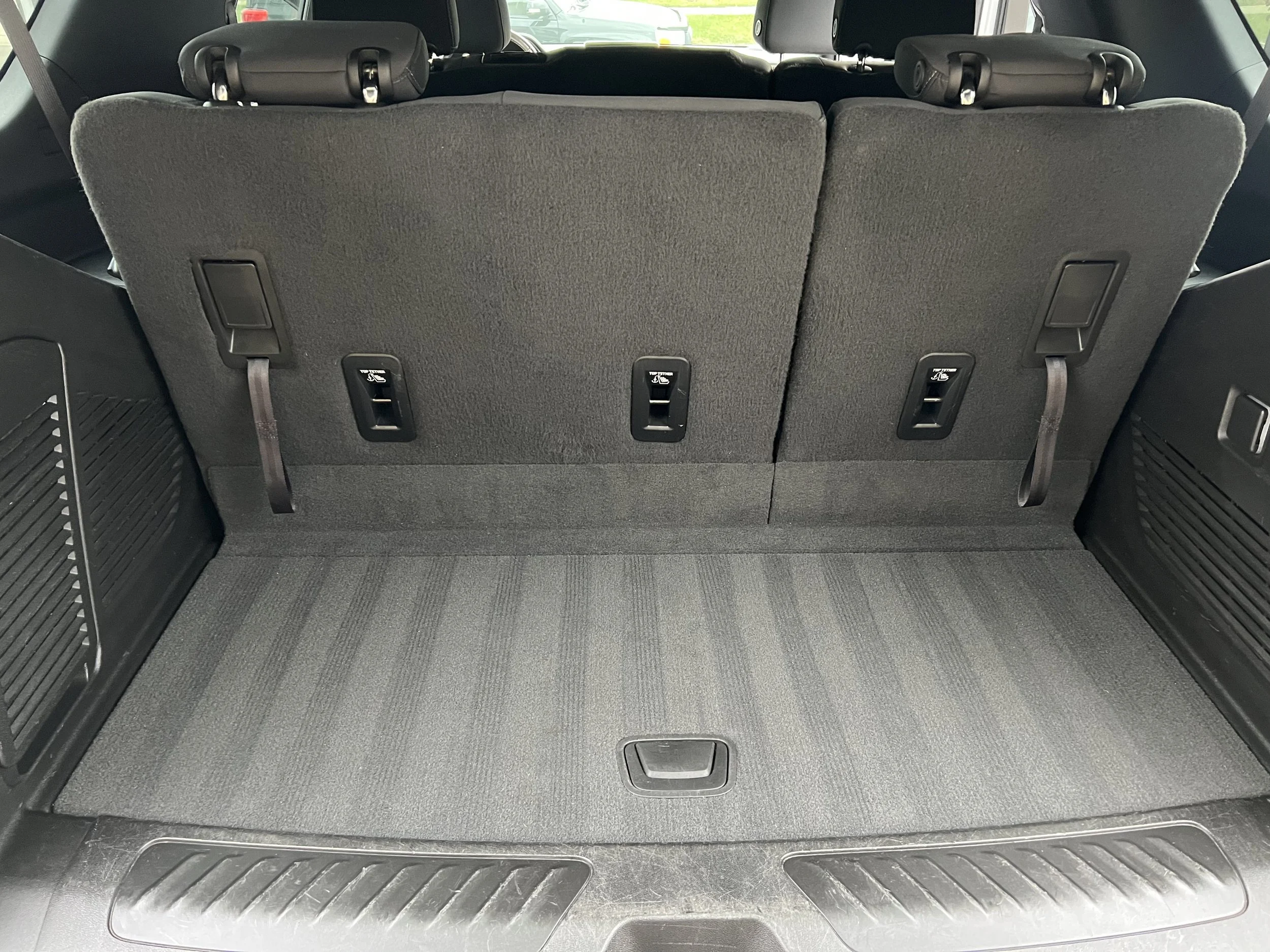 The image shows the cargo area of an SUV with the rear seats folded down, revealing an empty black carpeted space with seat release latches and hooks on the back of the seatbacks.