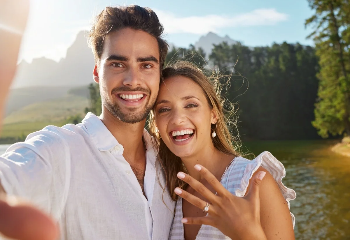Smiling couple taking a selfie outdoors; the woman shows an engagement ring with a lake and trees in the background.