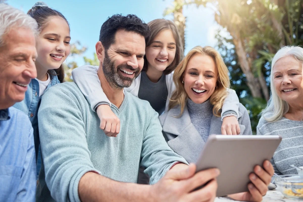 Multi-generational family smiling together outdoors while looking at a tablet, sharing a joyful moment