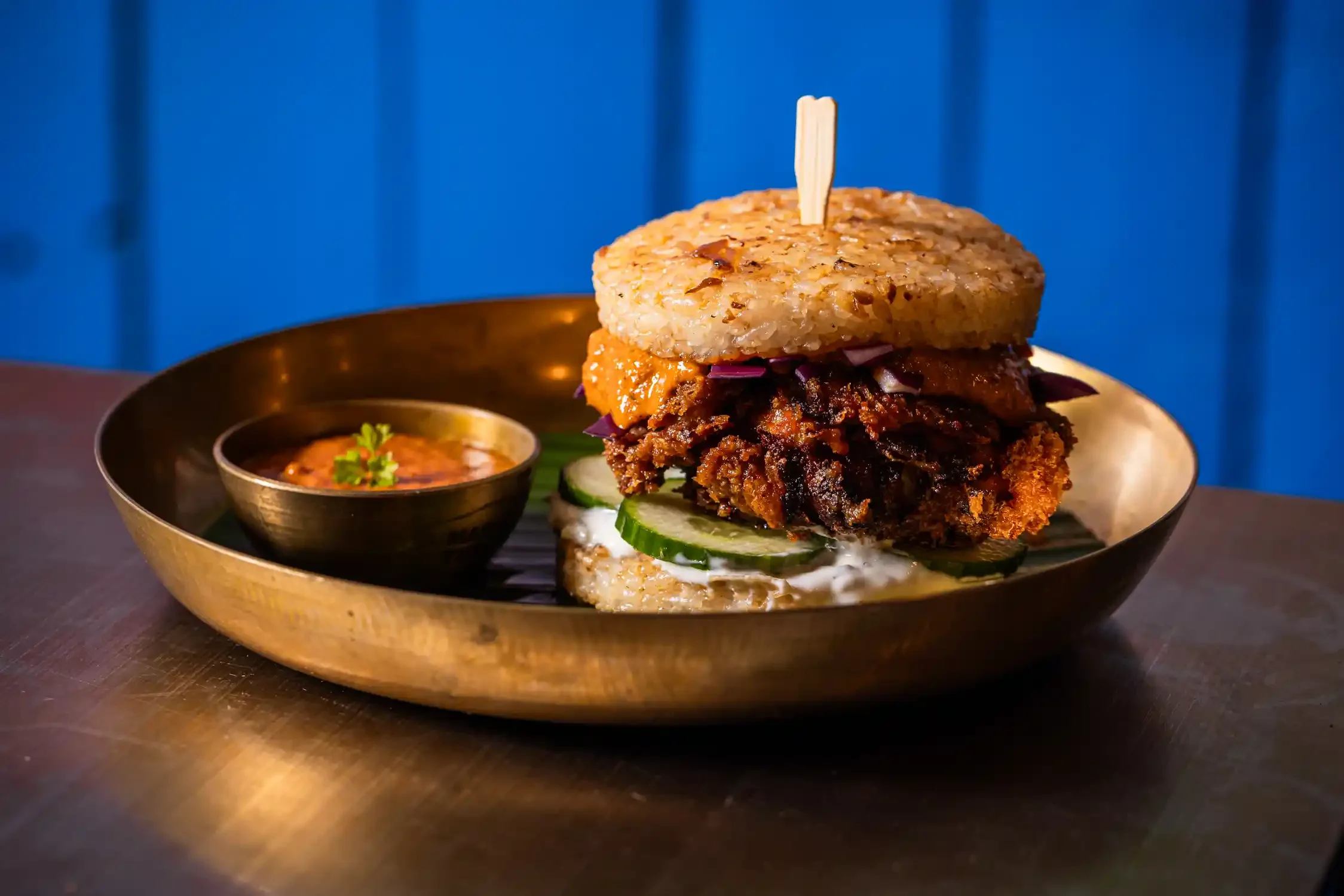 A burger with rice patties, fried chicken, cucumber slices, and purple cabbage, served with a side of chutney.