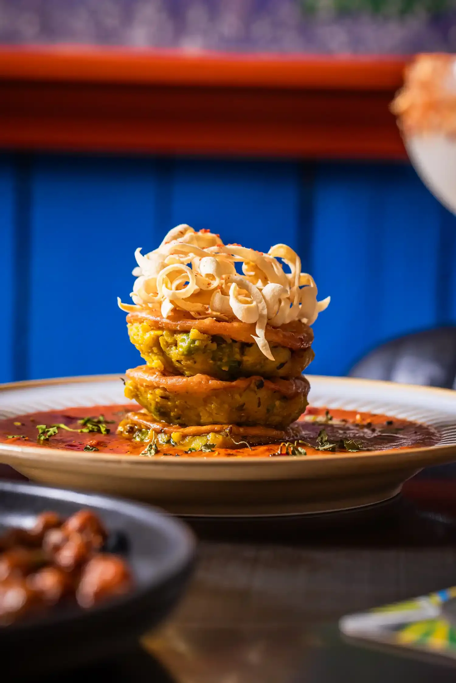 Stack of three veggie fritters topped with a portion of curly fried onions, served on a plate with sauce and garnished with herbs.