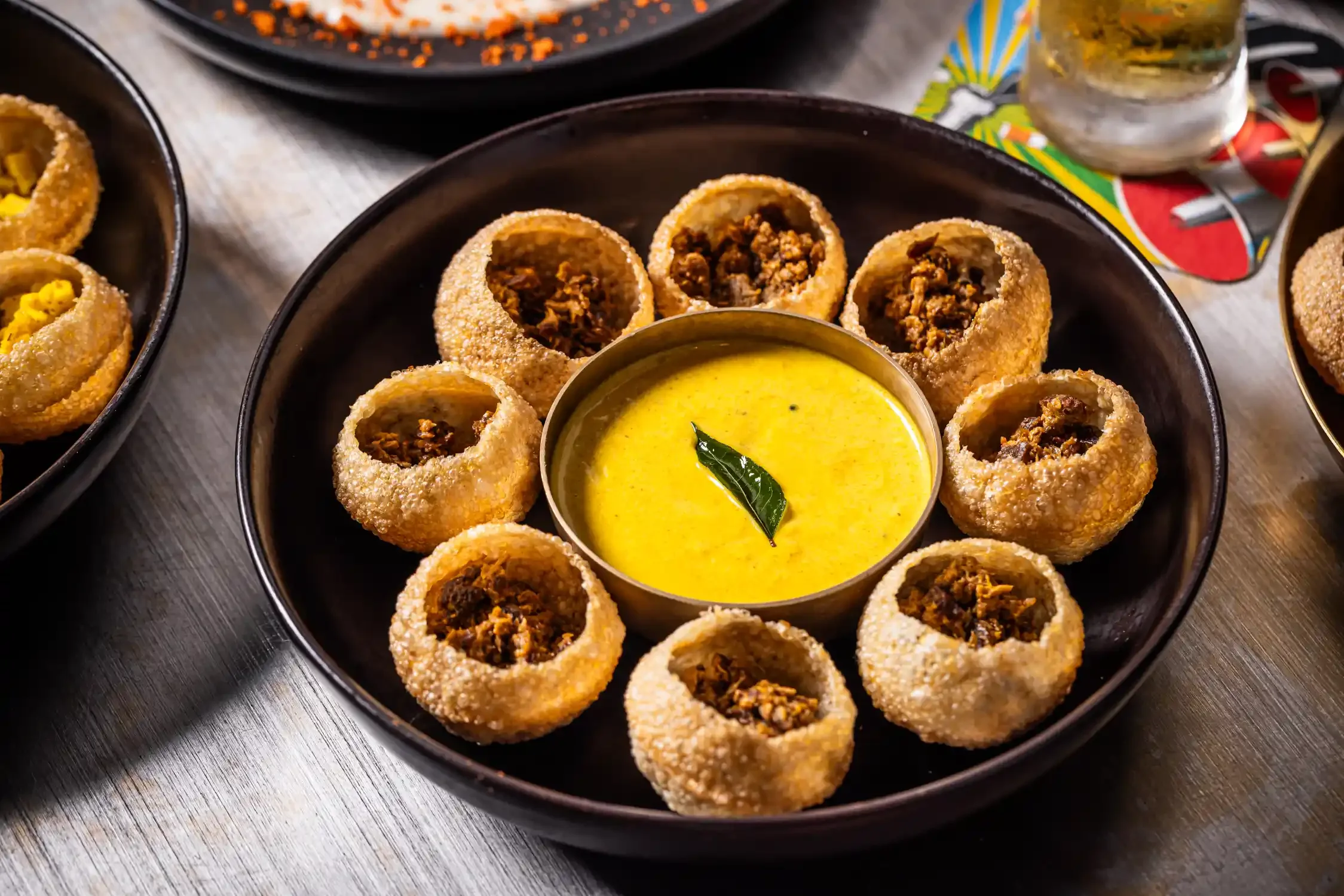 A black plate with ten crispy, fried Indian samosas filled with minced meat, arranged around a small bowl of yellow curry garnished with a green leaf.
