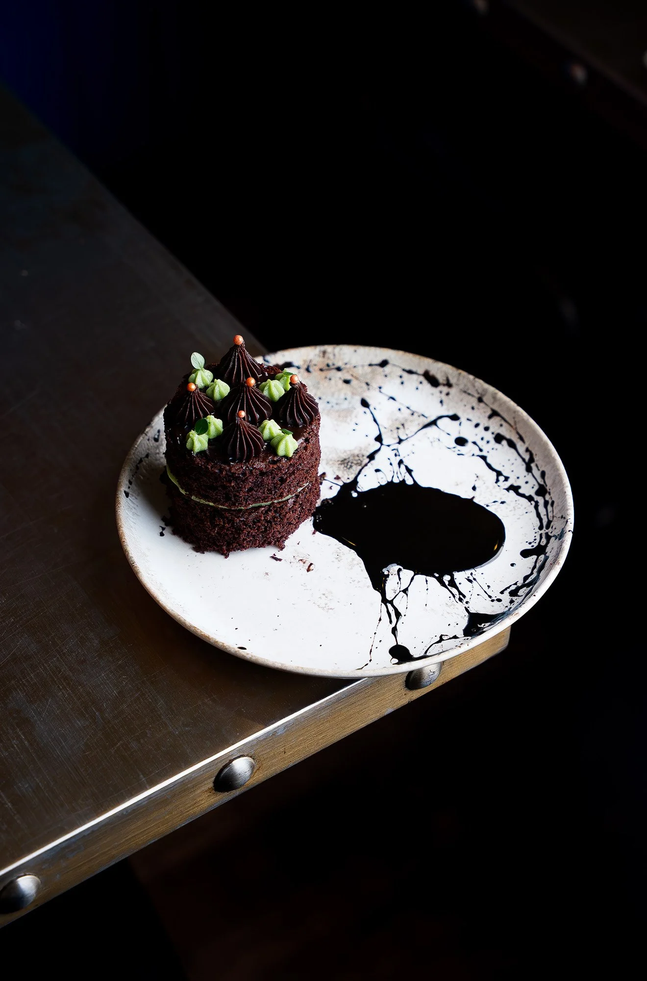 A slice of chocolate cake with dark frosting, topped with green and chocolate dollops, on a white plate with black sauce splatters, placed on a wooden table.