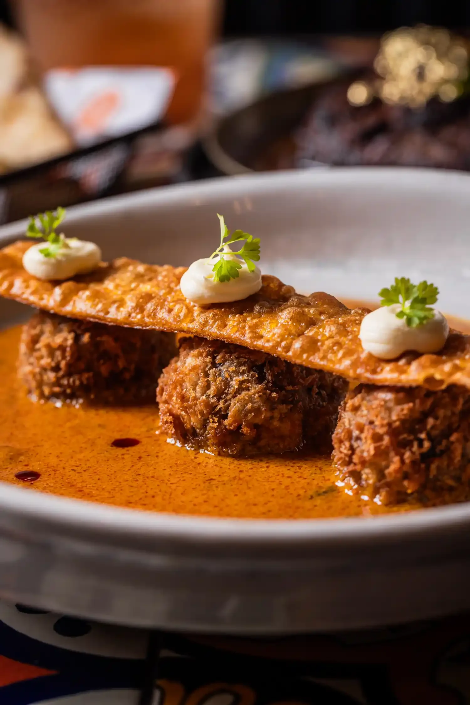 Close-up of a plate with three meatballs in sauce, topped with crispy cheese and garnished with small dollops of sour cream or similar, and tiny sprigs of parsley.