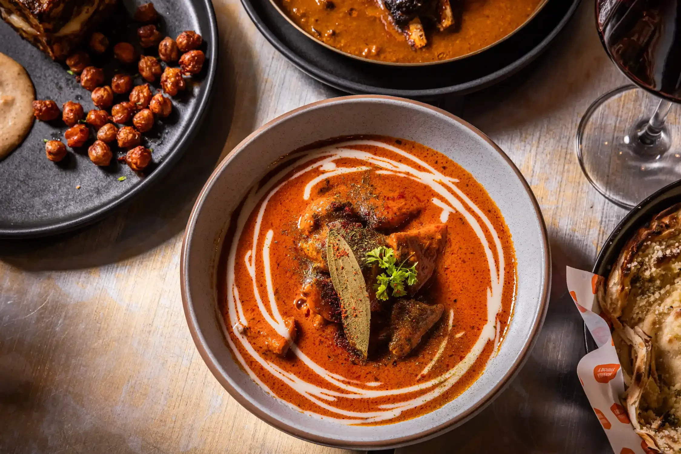 A bowl of Indian curry with a garnish of coriander, served with a side of roasted chickpeas and flatbread, and glasses of red wine.