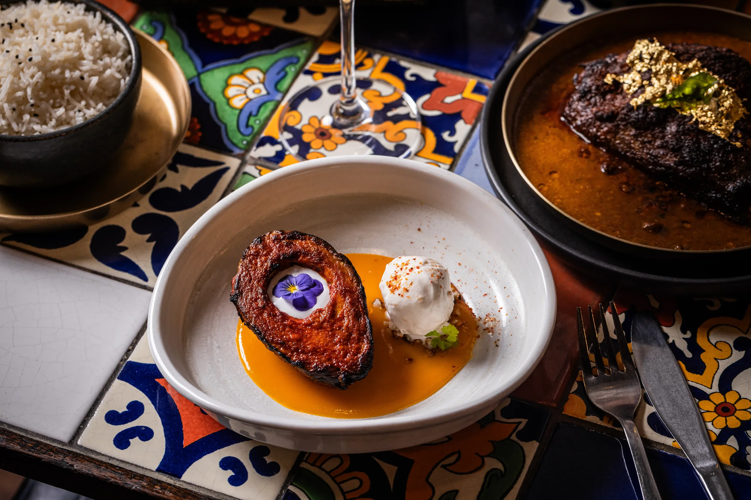 A plate with a slice of glazed meatloaf, a scoop of mashed potatoes with red pepper flakes, and an edible flower on top. Nearby are bowls of white rice and a braised meat dish with sauce and herbs on top. The setting features colorful, patterned tile