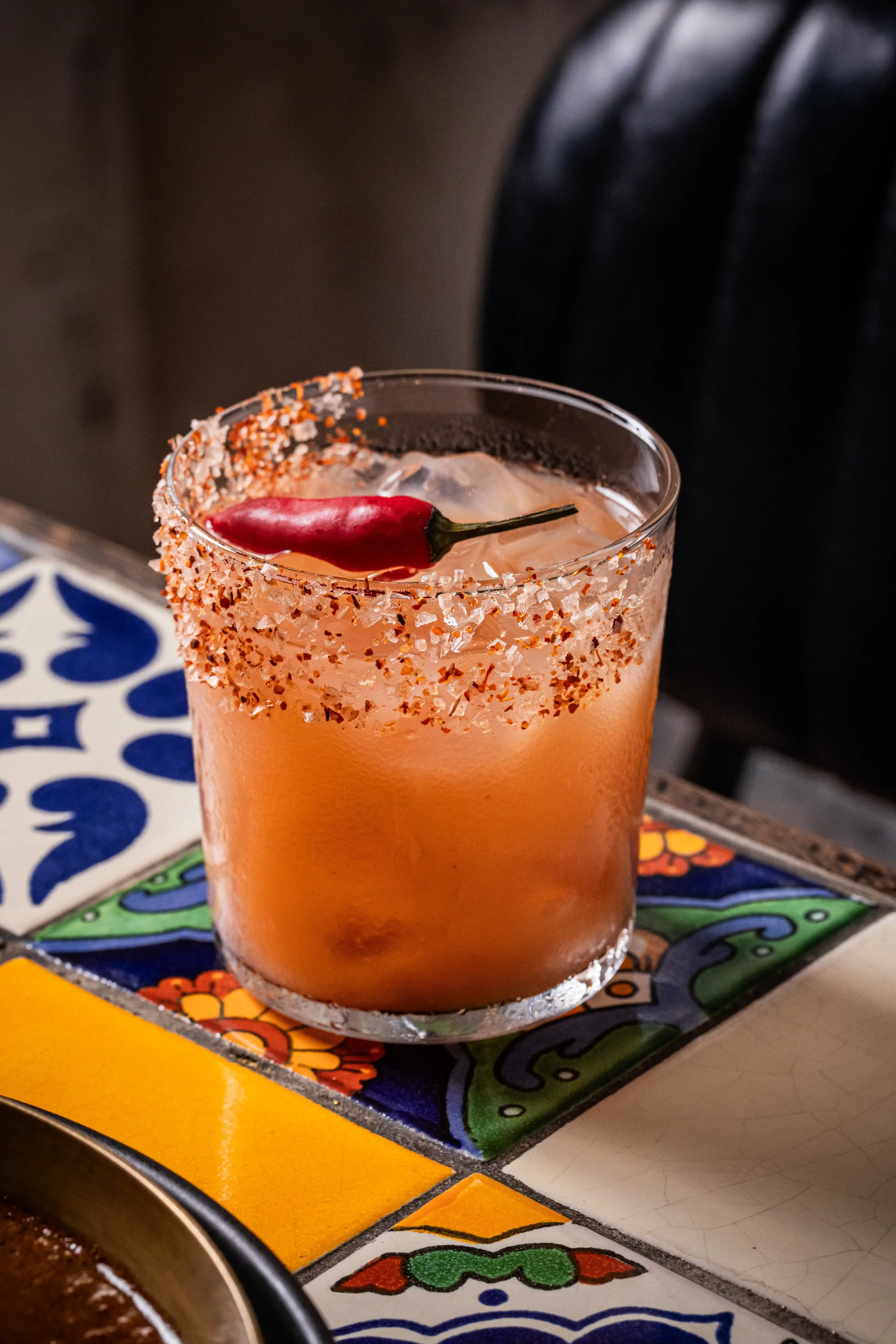 A pink cocktail with a red chili pepper garnish, salt and chili powder rim, served on a colorful tiled table.