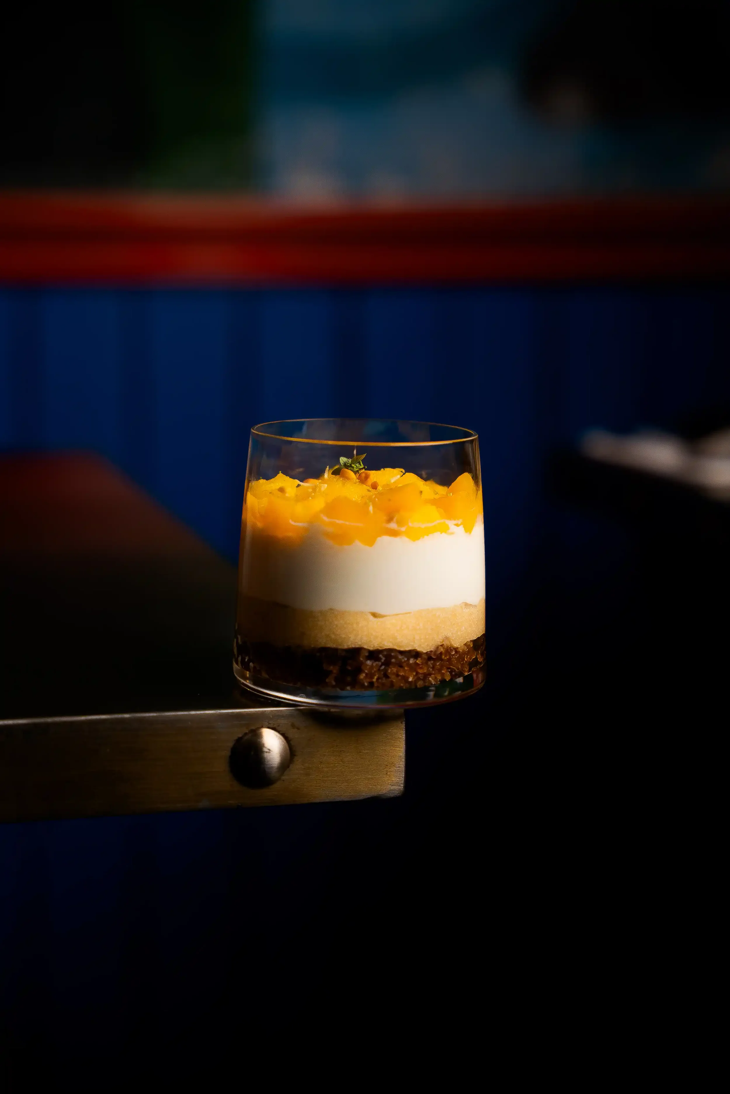 A layered dessert in a glass with a crumb base, creamy middle layer, and yellow fruit topping, garnished with a small green leaf.