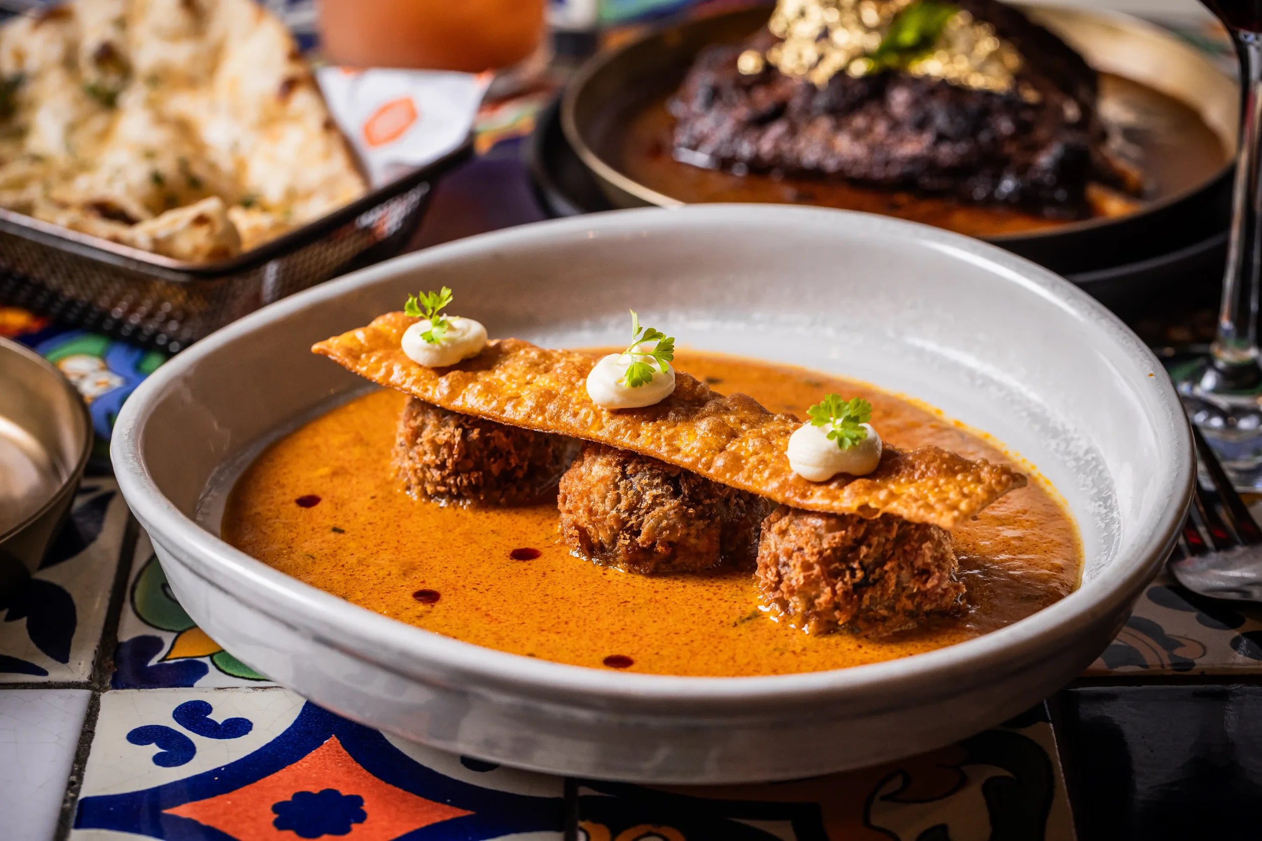A bowl of Mexican dish featuring meat in a red sauce, topped with a crispy tortilla strip, dollops of sour cream, and parsley, on a colorful tiled table with additional dishes and drinks in the background.