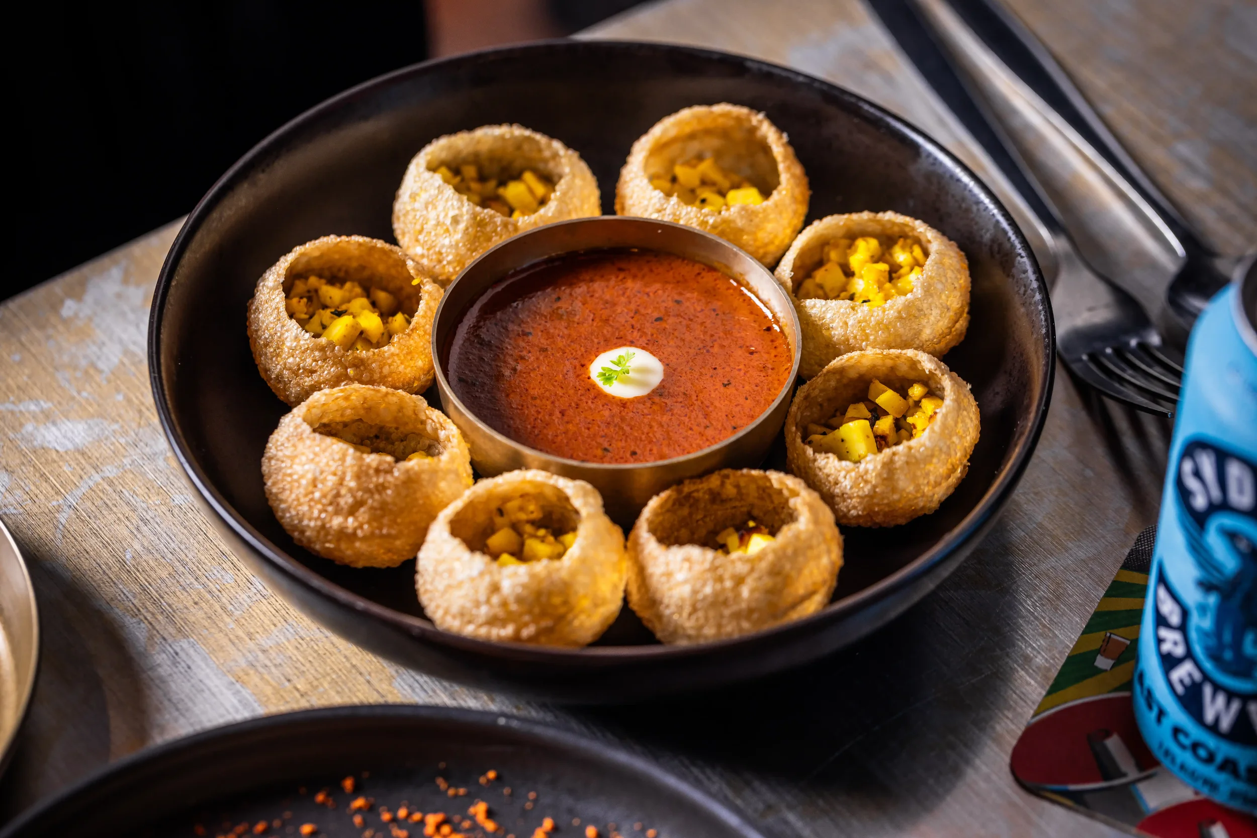 A black plate containing ten breaded and deep-fried jalapeño poppers filled with chopped yellow peppers, arranged in a circle around a small bowl of red dipping sauce with a dollop of white sour cream and a small sprig of cilantro in the center, on a