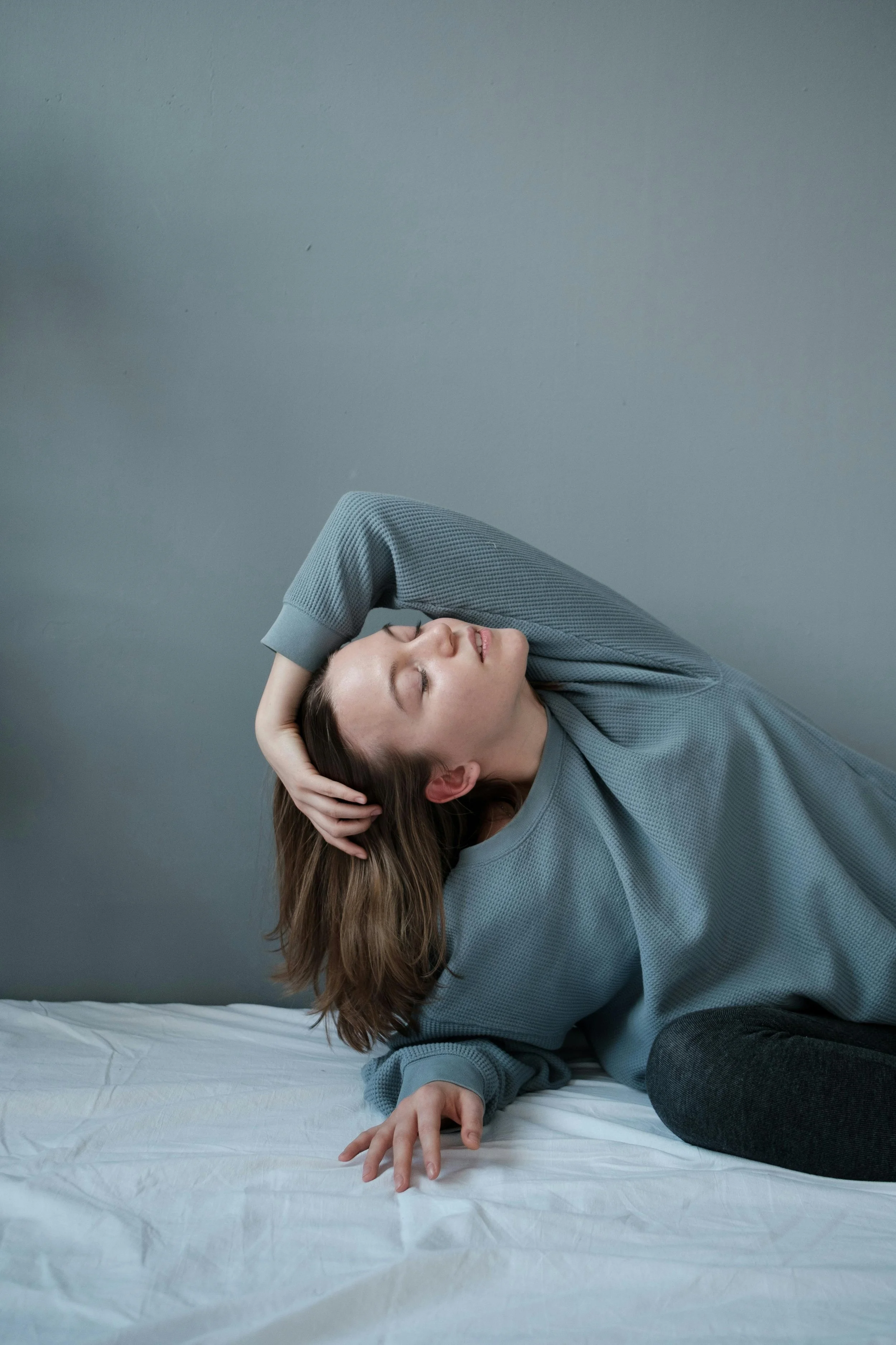A woman with brown hair wearing a gray long-sleeve shirt, lying sideways on a bed with a white sheet, touching her head with her left hand, and eyes closed.