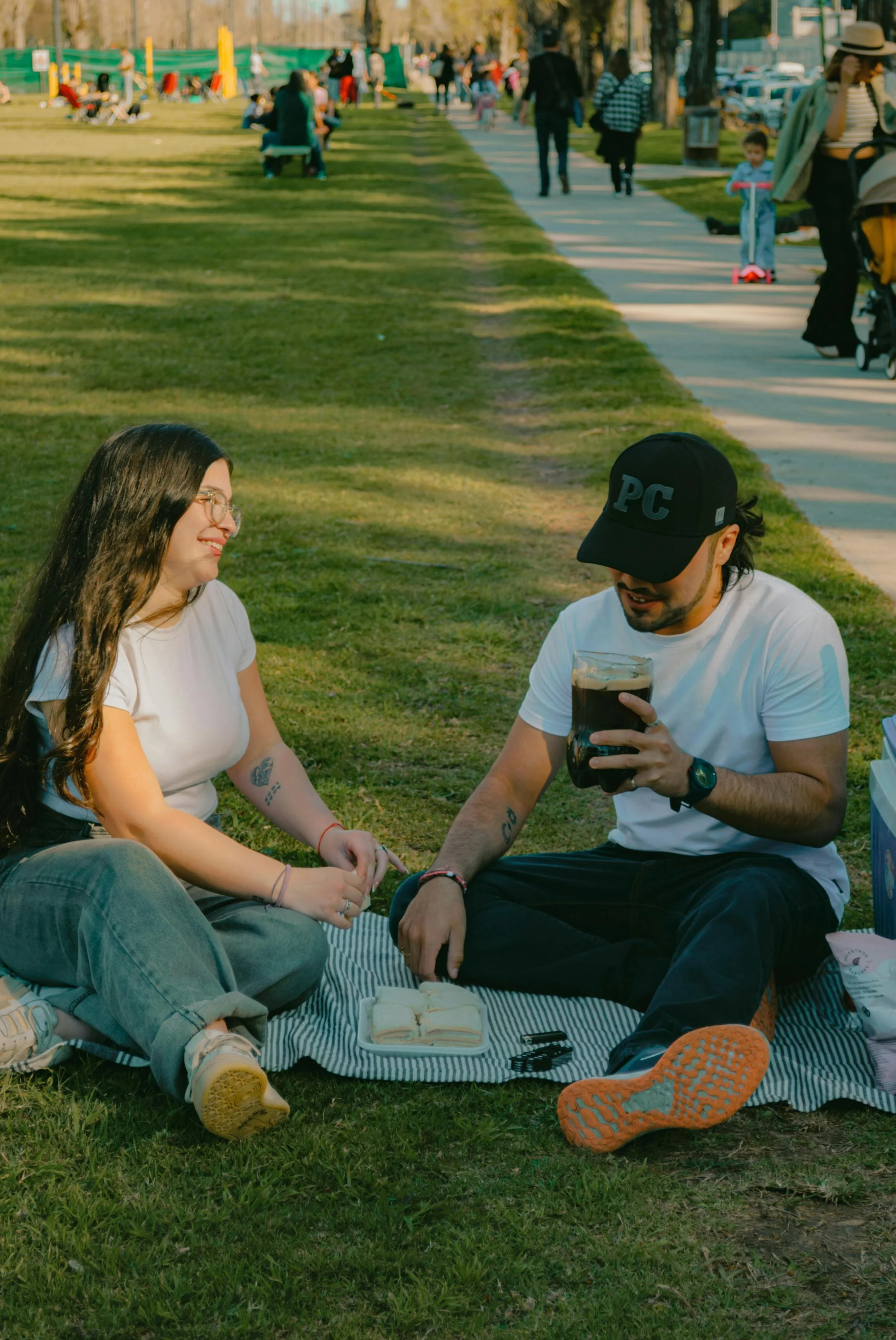 A woman and a man sitting on a picnic blanket outdoors in a park enjoying a picnic, with the woman smiling and the man holding a large drink. There are sandwiches on a tray between them and other park activity in the background.