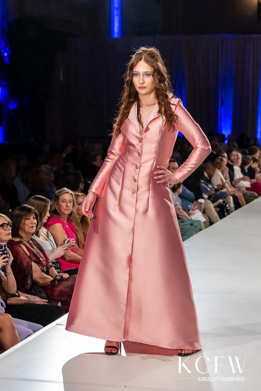 A female model walks on the runway at Kansas City Fashion Week, wearing a long, pink satin dress with buttons down the front and black high heels, while an audience watches.