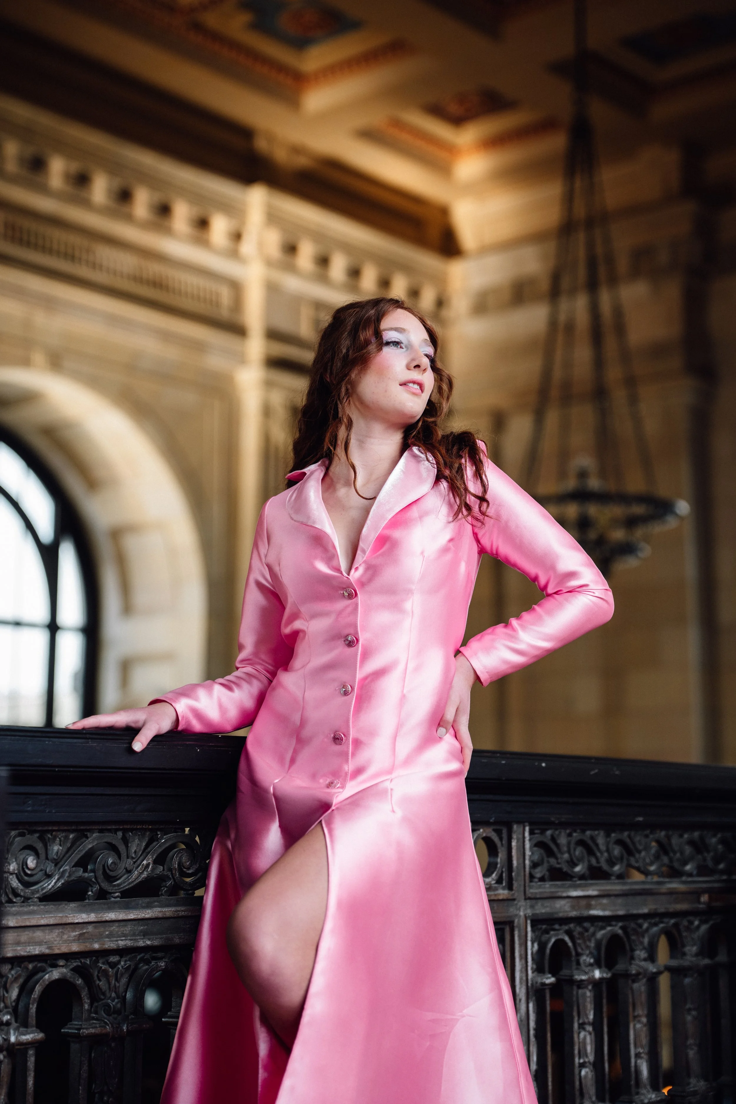 A woman in a shiny pink satin dress with a high slit, posing indoors near an ornate black railing with a large arched window and decorative woodwork in the background.