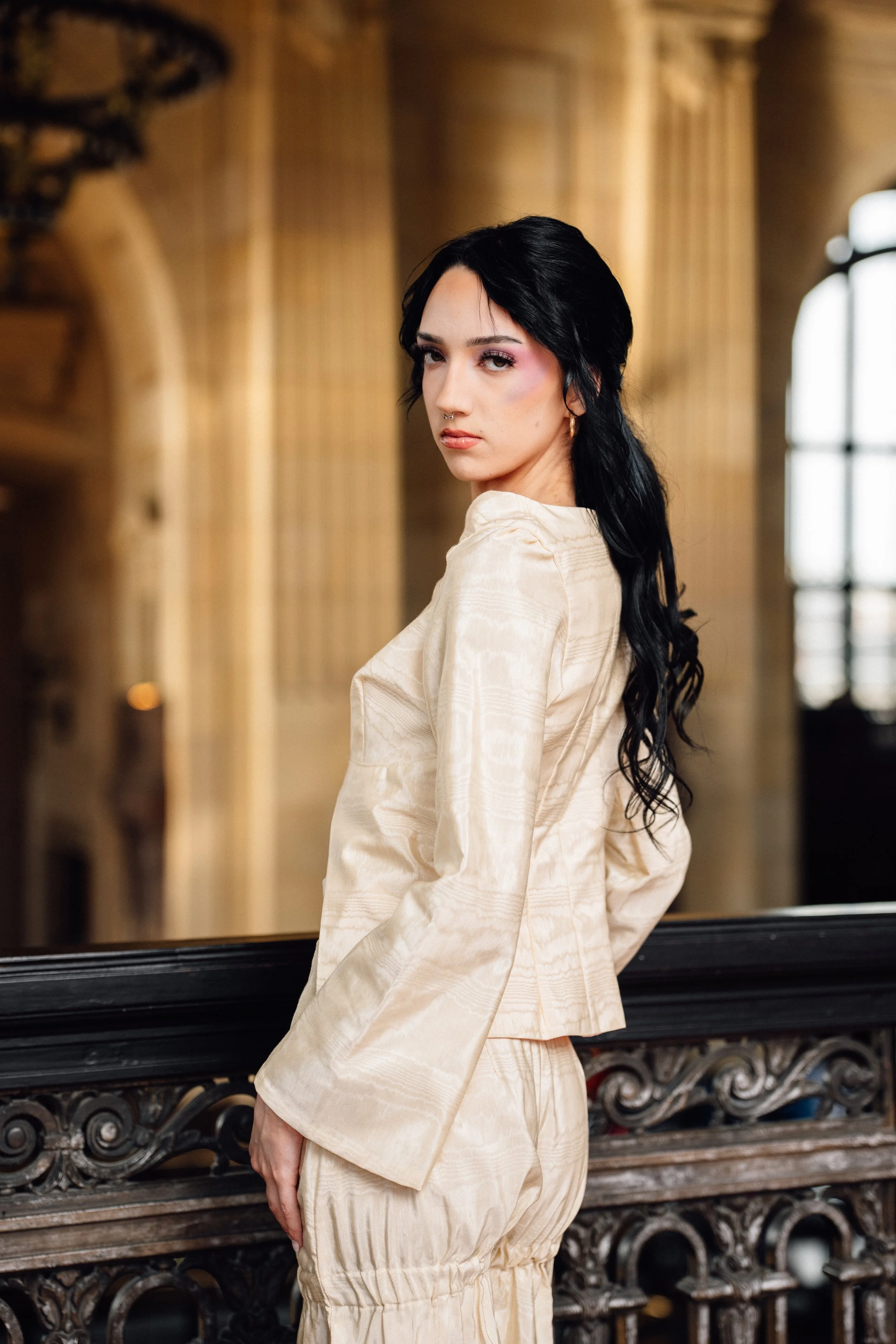 A young woman with long black hair, wearing a beige satin long-sleeve top and matching pants, standing indoors next to an ornate wooden railing, with a large window and classical architecture in the background.