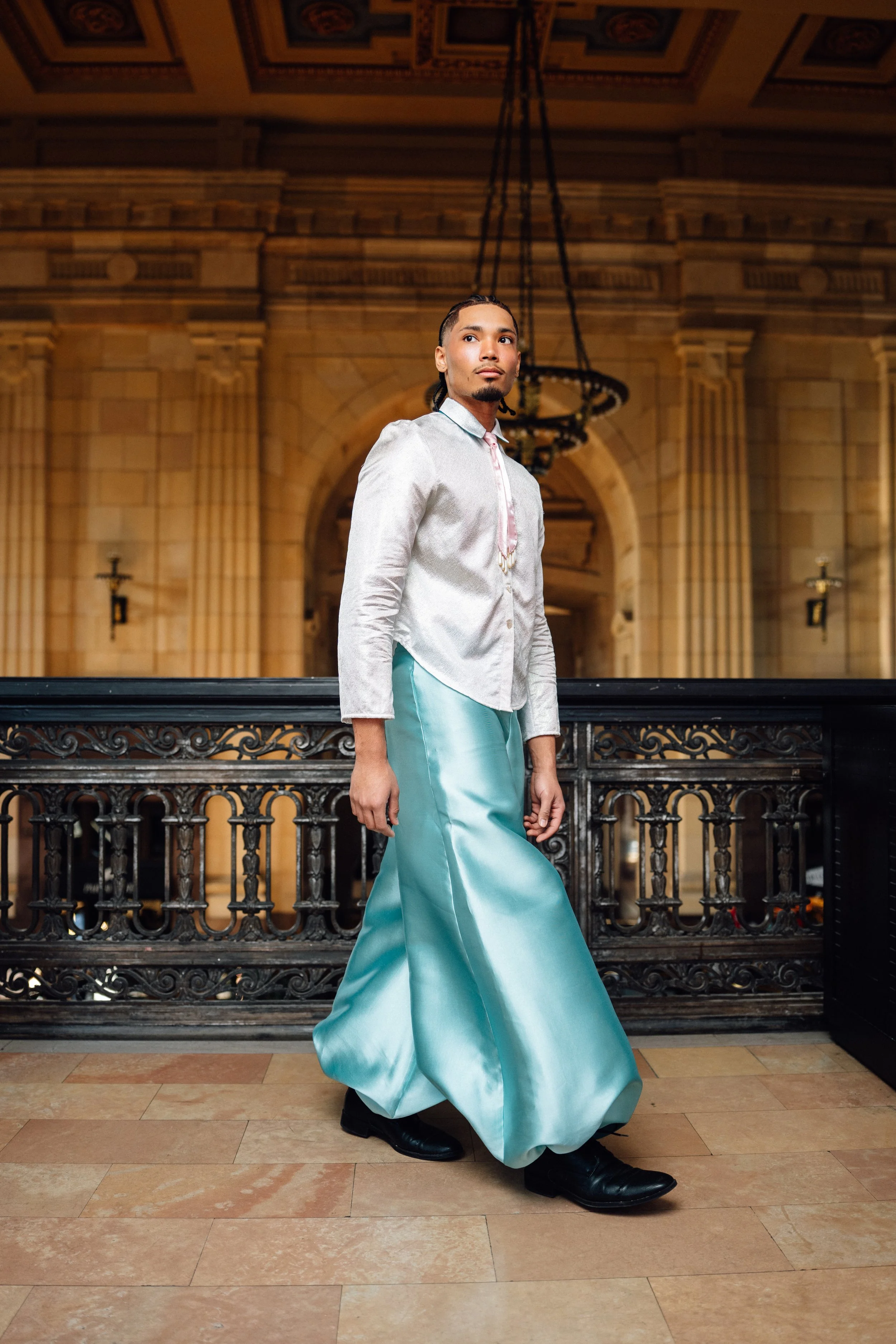 A man wearing a shiny silver shirt and wide teal satin pants standing in an elegant, historic building with ornate wall and ceiling details.