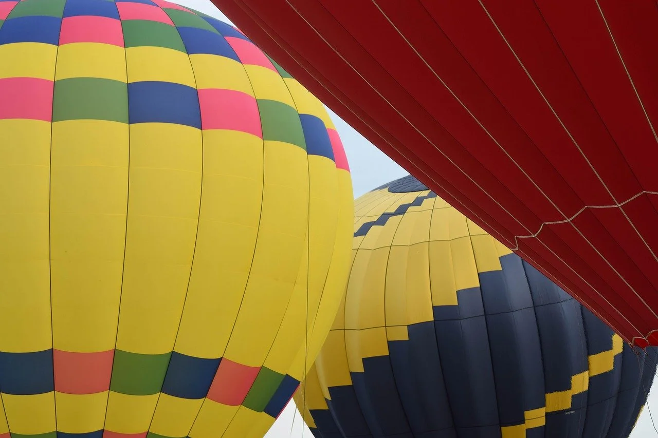 Colorful hot air balloons with yellow, red, blue, and green patterns, viewed from below against a light sky.