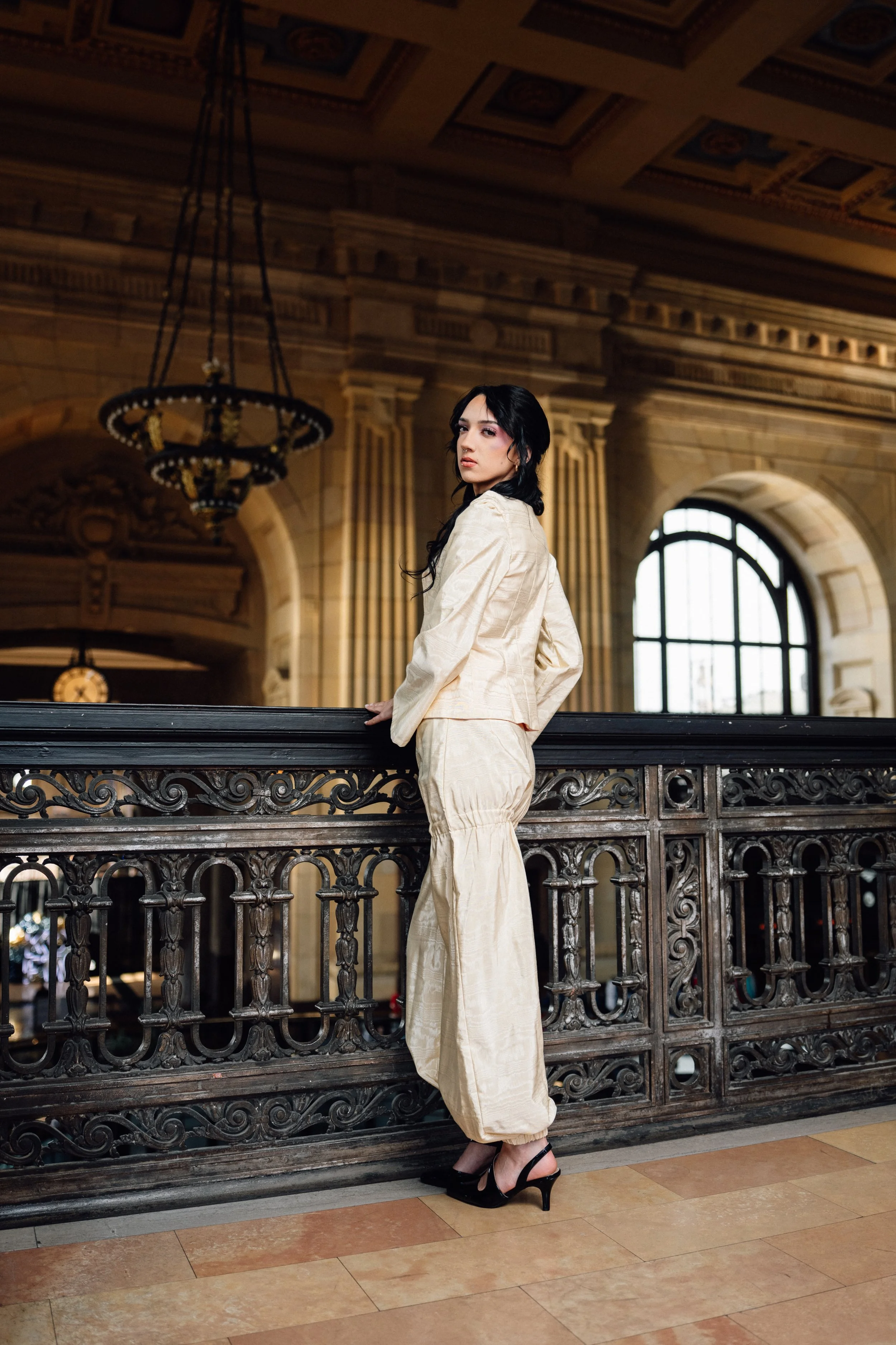 A woman with black hair in an elegant cream-colored suit and black heels standing in front of an ornate railing inside a grand historic building with high arched windows and a chandelier.