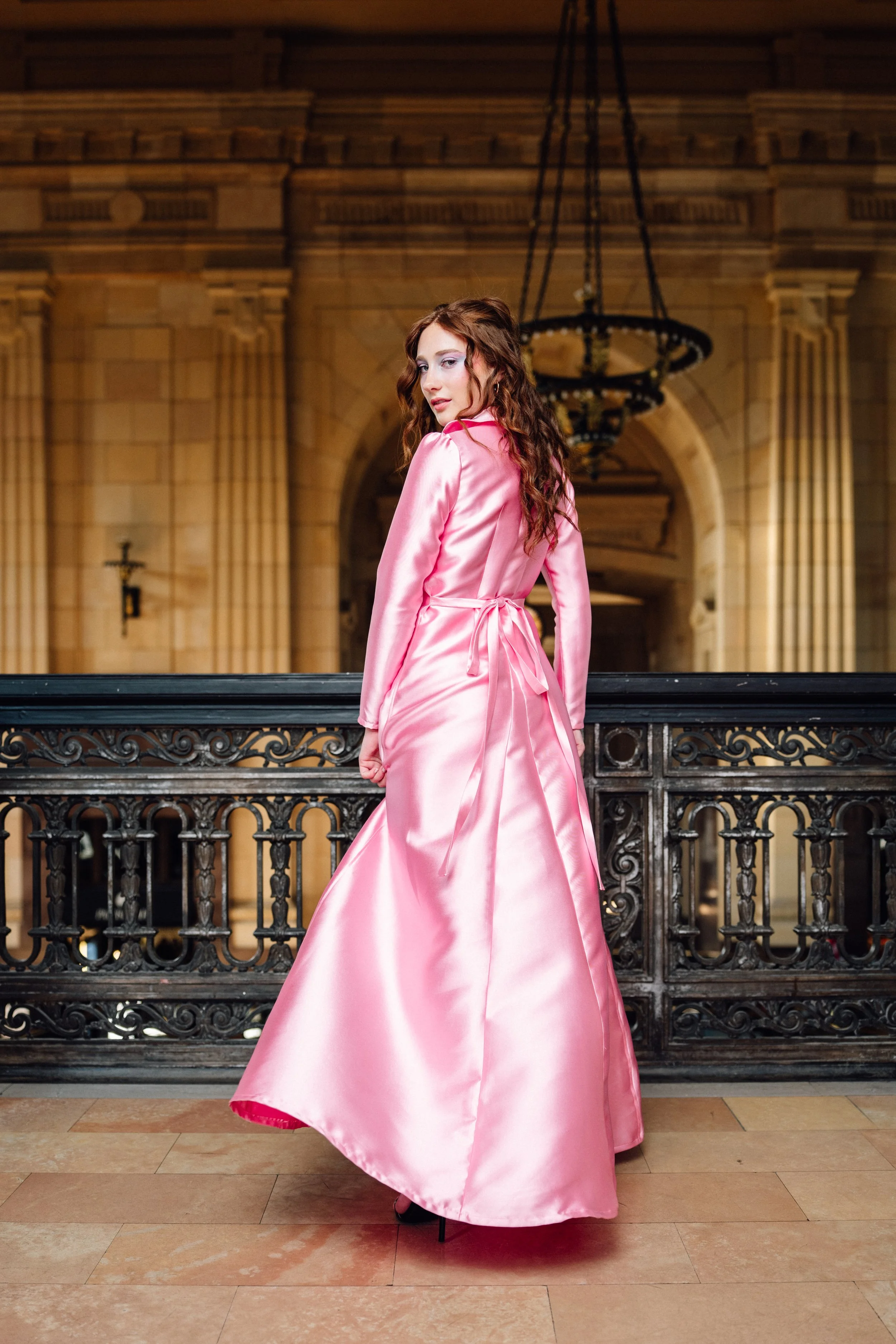 A woman in a pink satin gown stands on a balcony in an ornate hall with architecture featuring arches and a large chandelier.