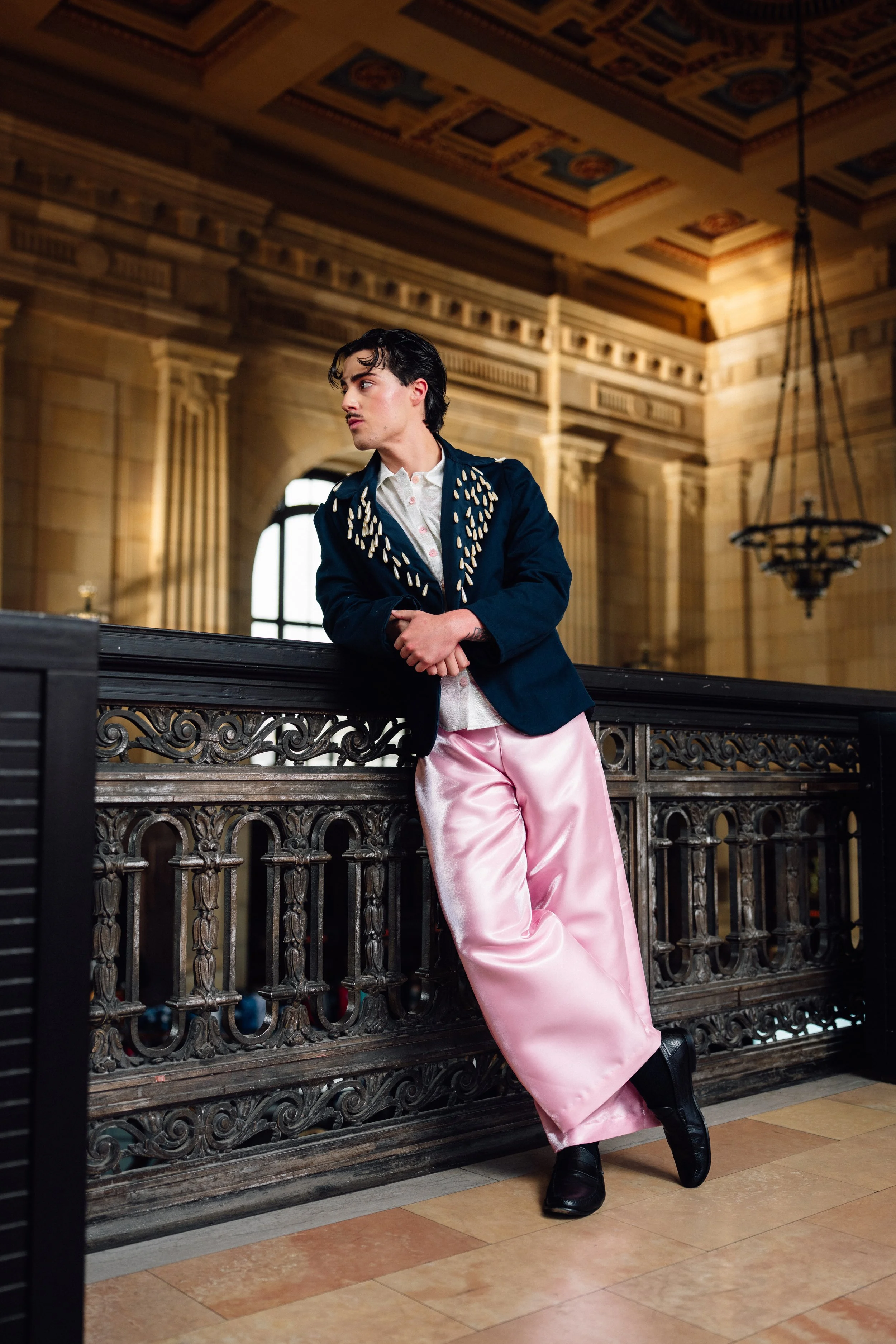 A young man leans on an ornate wooden railing inside a grand, historic building with high ceilings, decorative ceiling details, and large windows. Model wearing Sarah Skelly Dreamscape outfit in studio lookbook photo