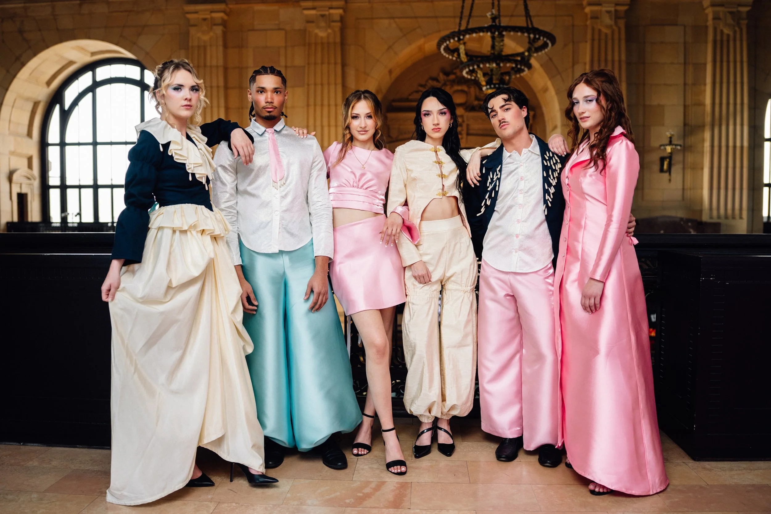 A group of six young people dressed in fashionable, vintage-inspired clothing, standing inside a grand, historic building with large arched windows and ornate chandeliers.