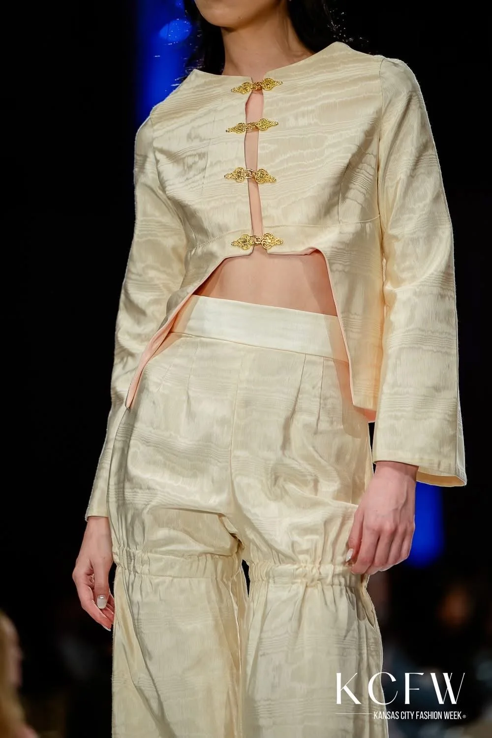 A model walking on a runway at Kansas City Fashion Week, wearing a cream-colored, satin-like, two-piece outfit with a cropped jacket fastened with gold decorative clasps and matching loose-fitting pants with gathered ankles.