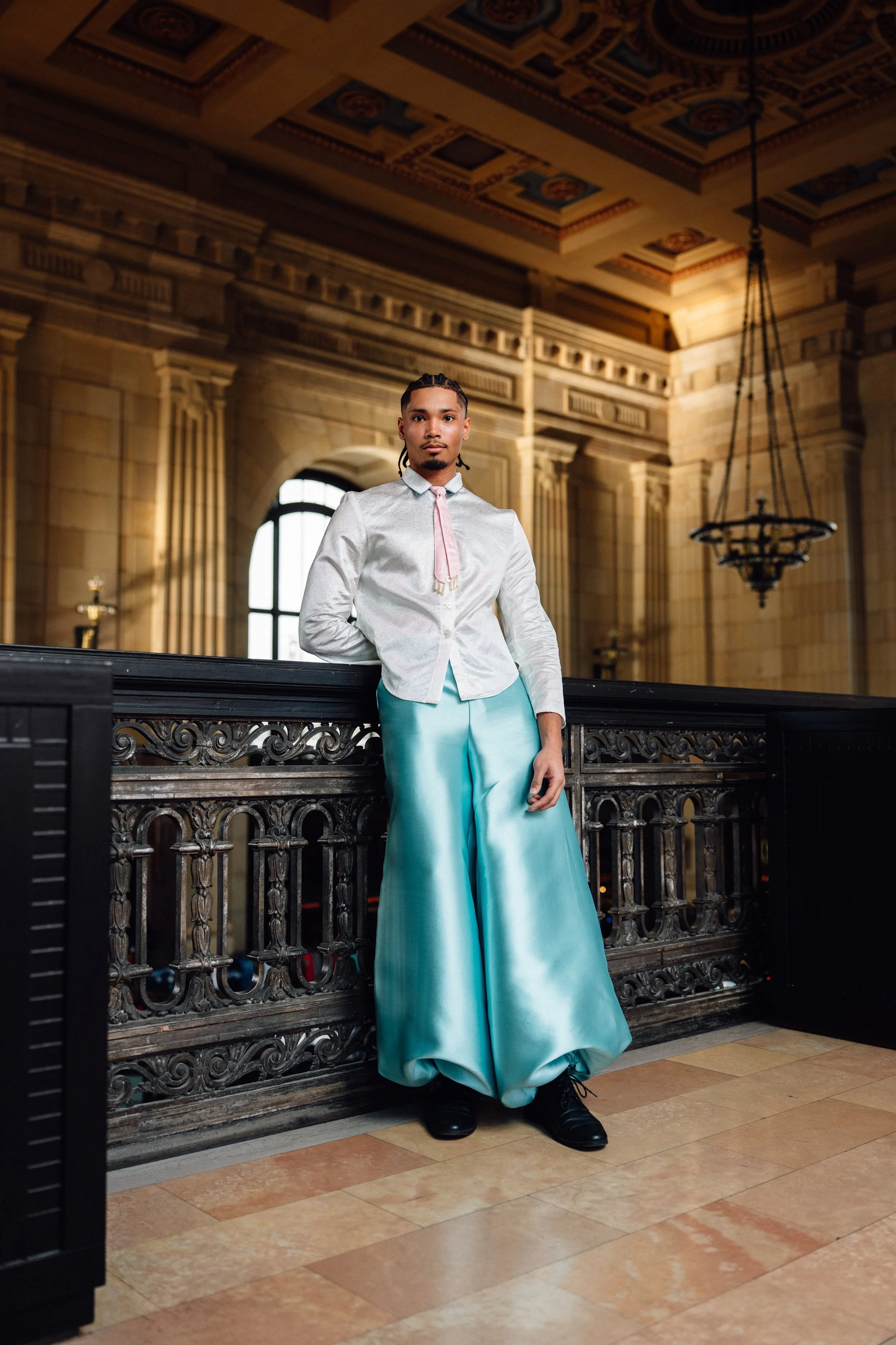 A man stands in a historic, ornate building with classical architecture, large windows, and elaborate ceiling details. He wears a white shirt, pink tie, and long, wide, silk turquoise pants.