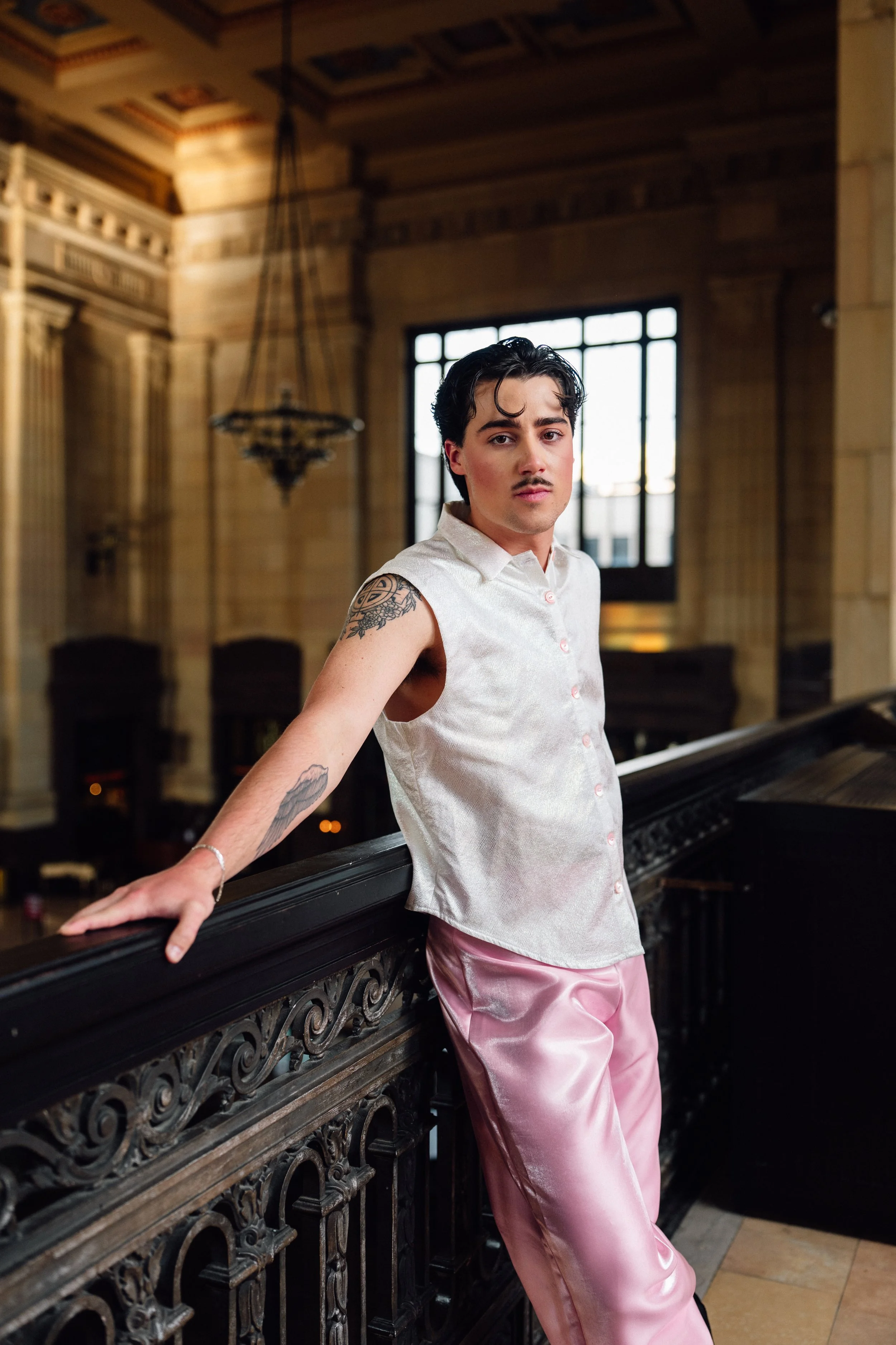 Young man with dark hair and tattoos wearing a sleeveless beige button-up shirt and pink satin pants leaning on a black ornate railing inside a grand, historic building with tall windows and wooden interior.