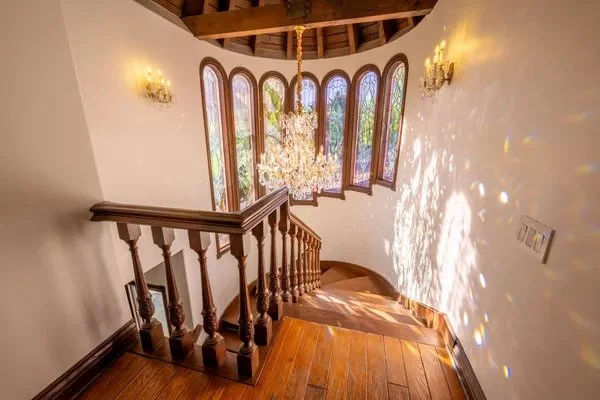 Interior view of a staircase landing with wooden stairs, a curved wooden railing, and large arched windows. A chandelier hangs from the ceiling, and sunlight creates patterns on the wall.
