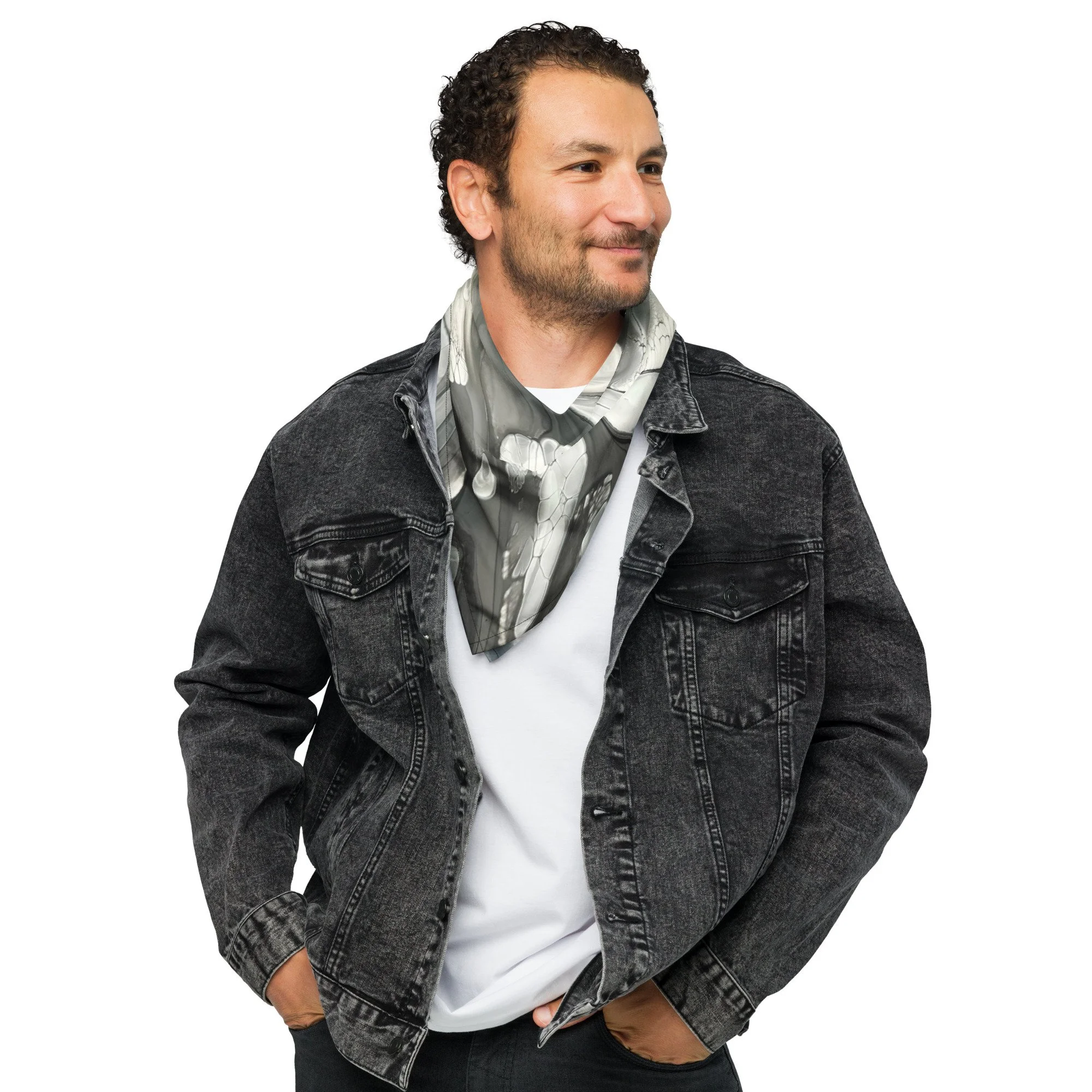 A man with curly hair and a beard wearing a black denim jacket, white t-shirt, and a gray patterned scarf, standing with hands in pockets and smiling.