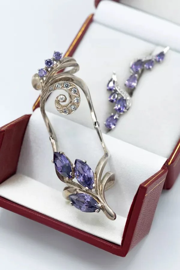 A jewelry set in a red box including a silver bracelet with purple gemstones and a matching ring and earrings, all decorated with lilac and clear stones.