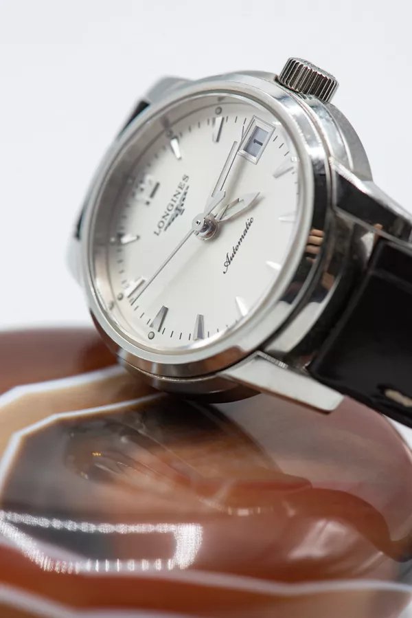Close-up of a silver Longines automatic wristwatch with a white dial, black leather strap, date display, and silver hour markers, resting on a polished wooden surface.