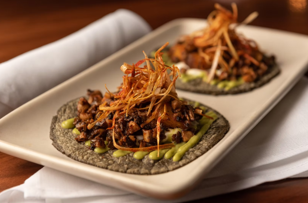 Two tacos on a rectangular white plate with a dark corn tortilla, filled with diced meat, topped with shredded fried vegetables, and drizzled with green sauce.