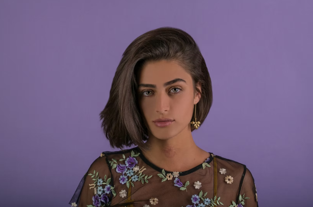 Portrait of a young woman with shoulder-length dark hair, wearing a sheer black top with colorful floral embroidery and a gold earring, against a purple background.